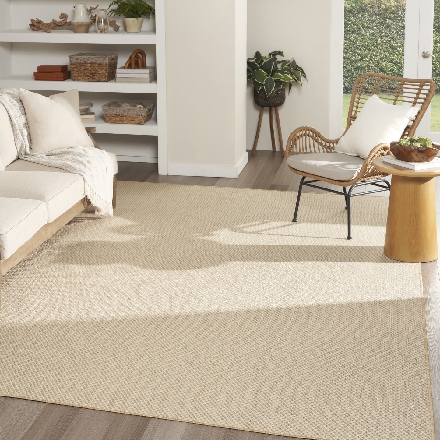 Bright living room with large beige textured rug, cream sofa, wicker lounge chair, round wooden side table, and potted plant.