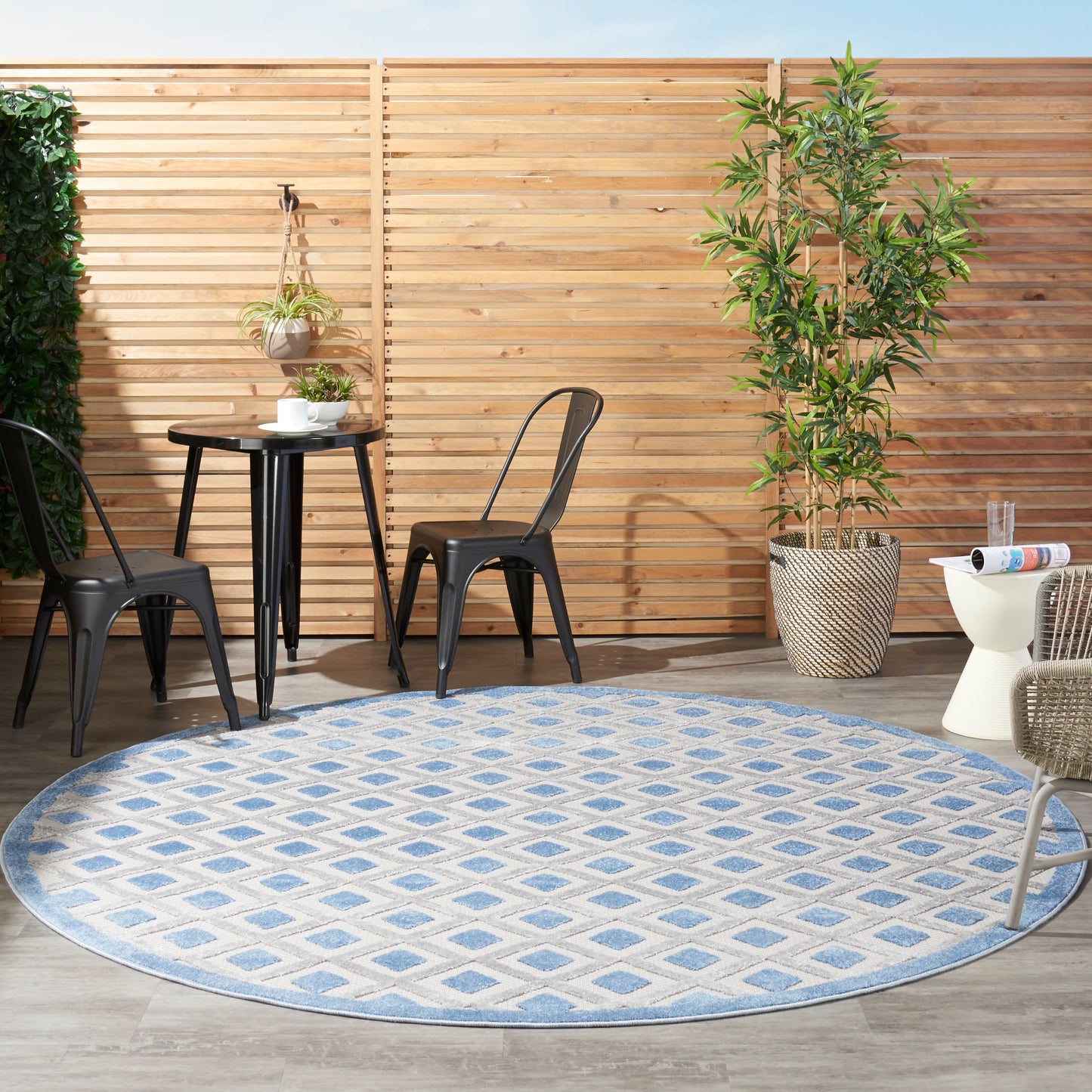 Outdoor patio with a round blue and white geometric patterned rug, black metal chairs, a small table, and a tall potted tree against a wooden fence.