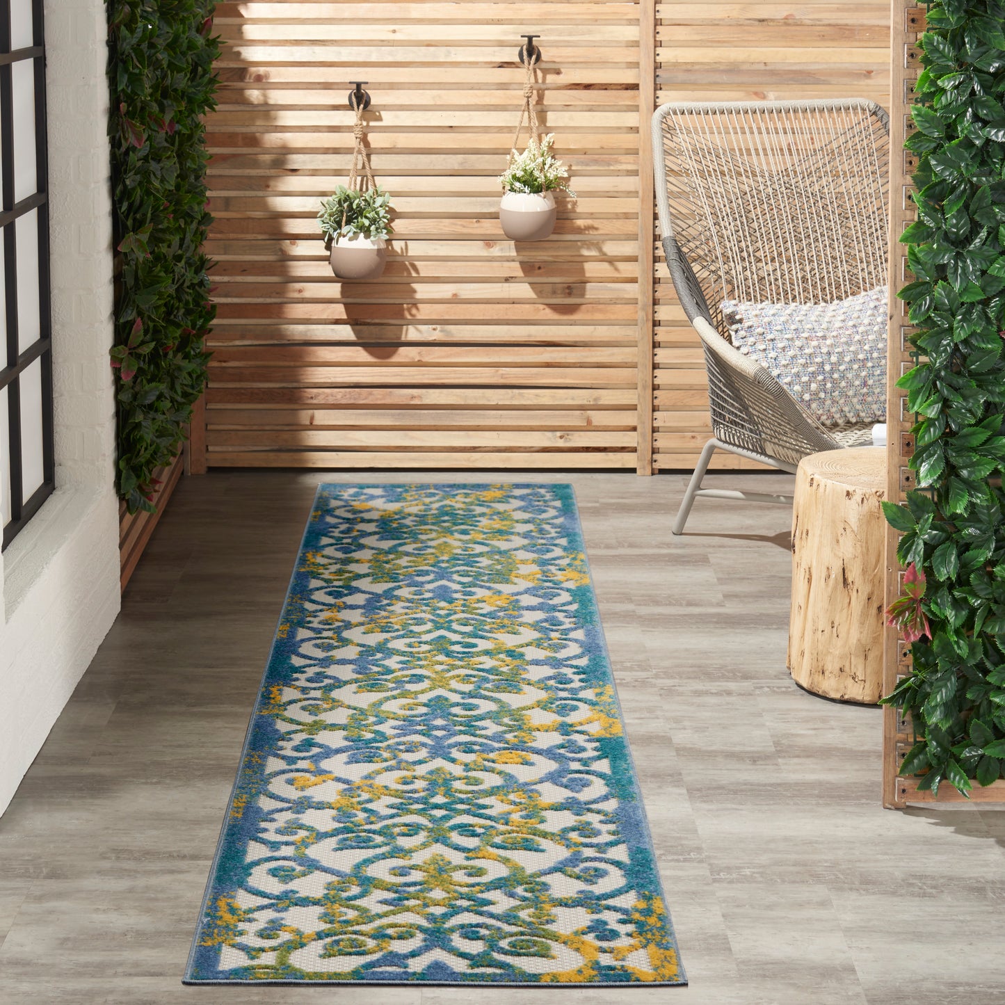 Long blue patterned outdoor rug on a gray wood floor beside a wicker chair, wooden slat screen and hanging planters.
