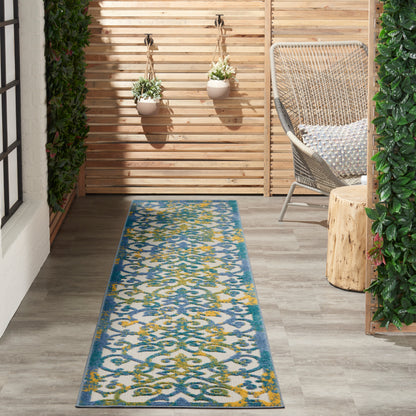 Long blue patterned outdoor rug on a gray wood floor beside a wicker chair, wooden slat screen and hanging planters.
