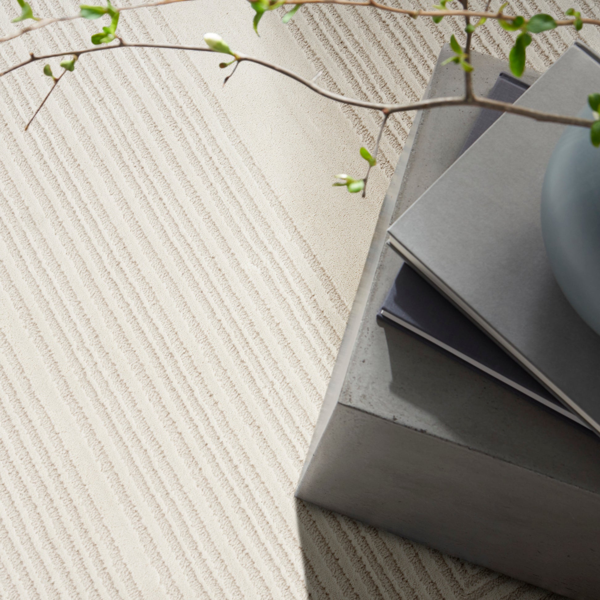 Beige striped rug beside a gray concrete block table with stacked notebooks and a pale ceramic vase, a trailing green plant.
