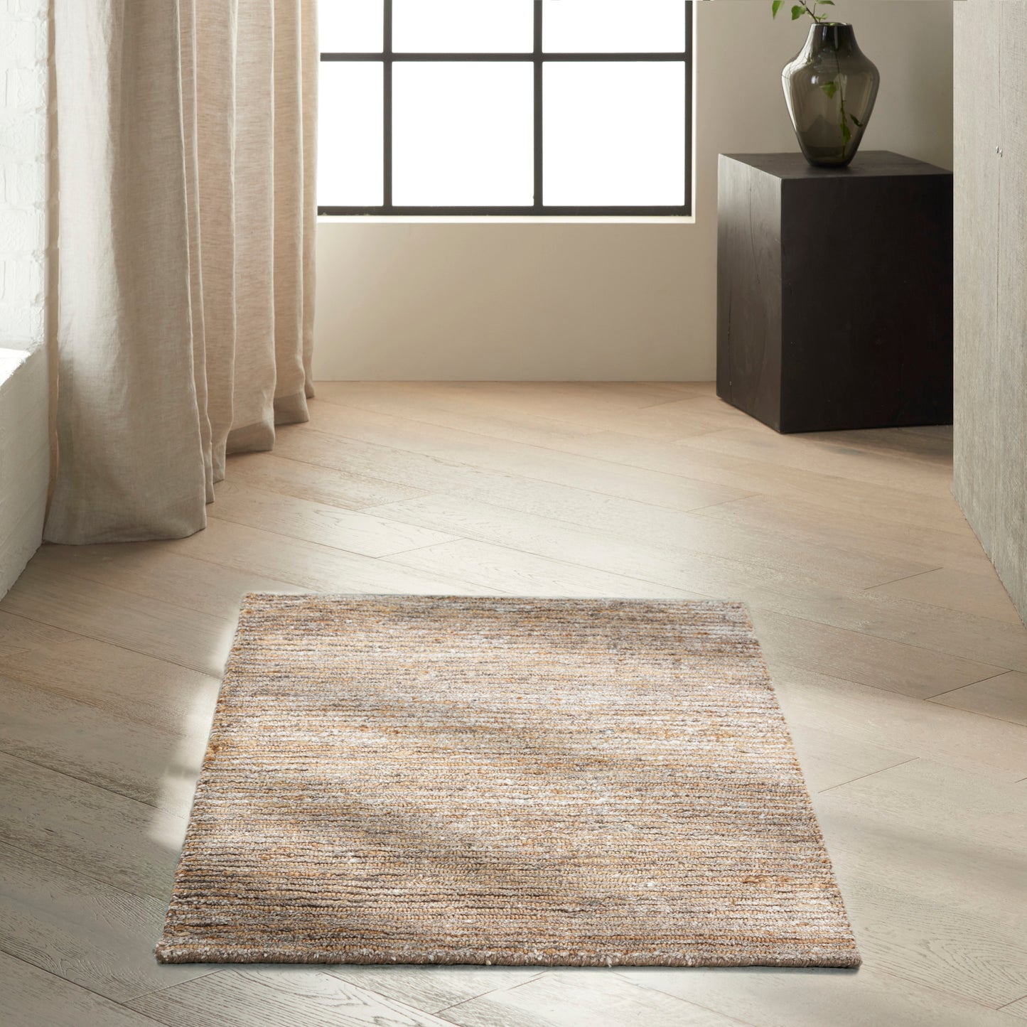 Beige textured rectangular rug on light wood floor in a bright minimalist room with sheer curtains and a dark pedestal with vase.