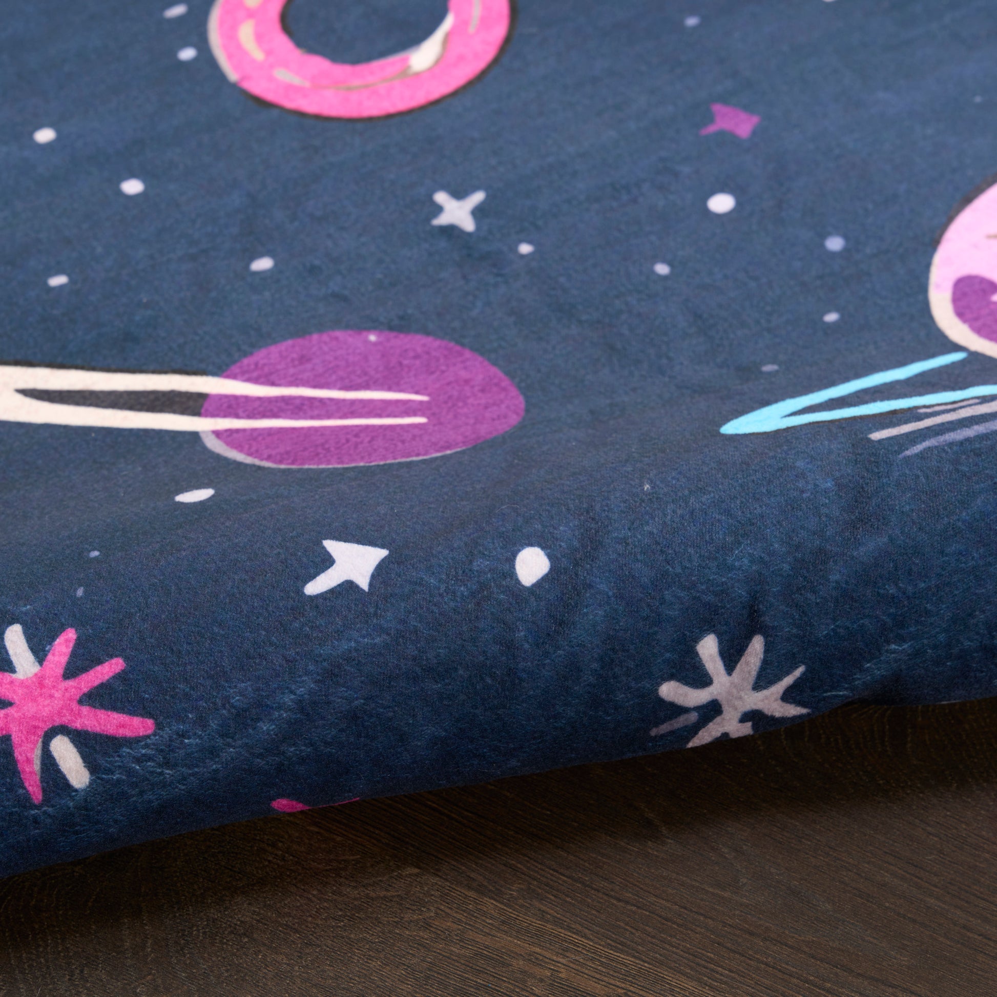 Navy blue space-print rug with pink and purple planets, stars and comets, folded edge on dark wood surface.