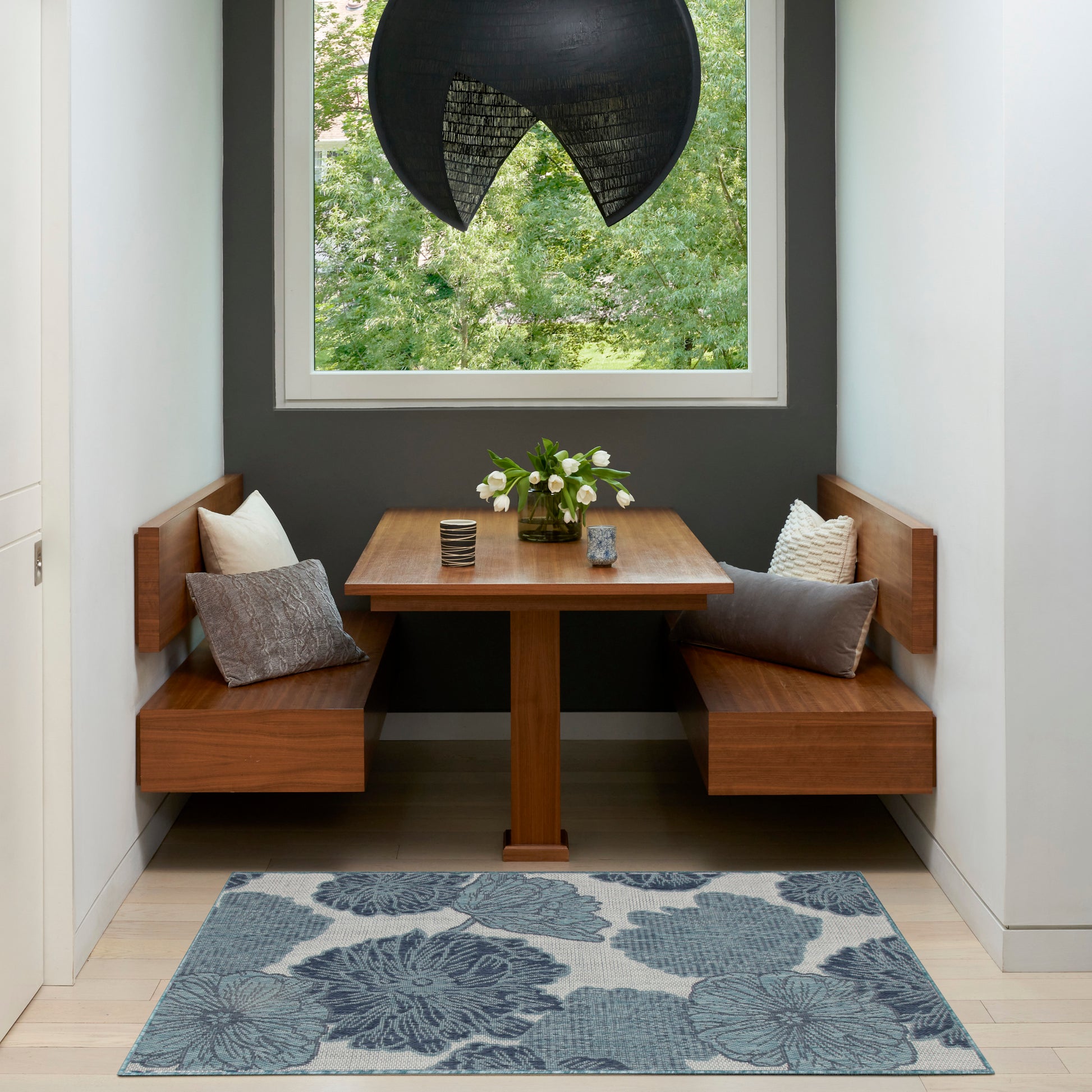 Warm wooden corner dining nook with built-in benches, square table, blue floral rug, white flowers, and black dome pendant.