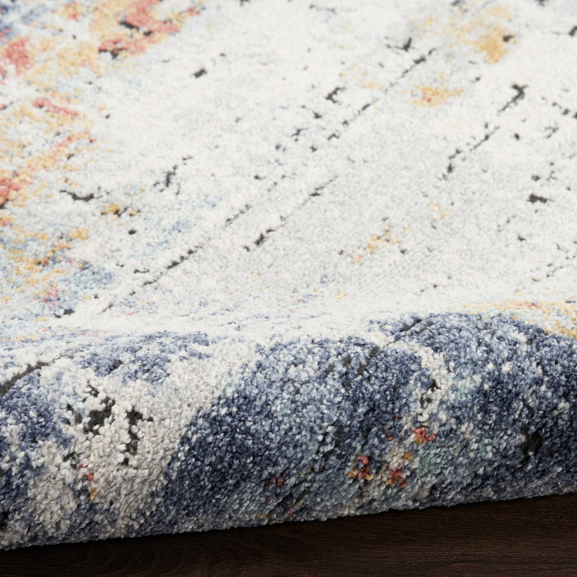 Close-up of a soft pile rug in cream and blue-gray with orange accents; rolled edge reveals an abstract pattern, ideal as an area rug.