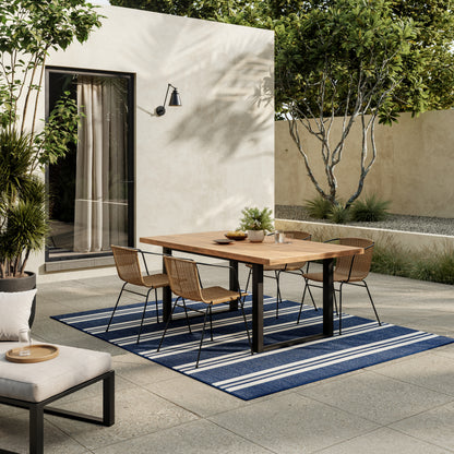 Outdoor dining set: wooden table with black legs and six woven chairs on a blue striped rug amid beige patio and greenery.
