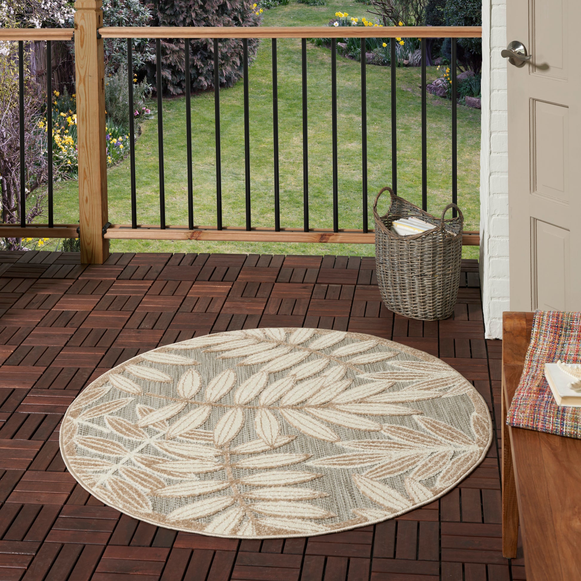 Round outdoor rug with beige and gray leaf pattern on a wooden deck, wicker basket nearby, porch door to the right.