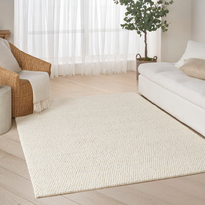 Cream textured rectangular area rug in a bright living room with white sofa, wicker chair, potted tree, light wood floors.