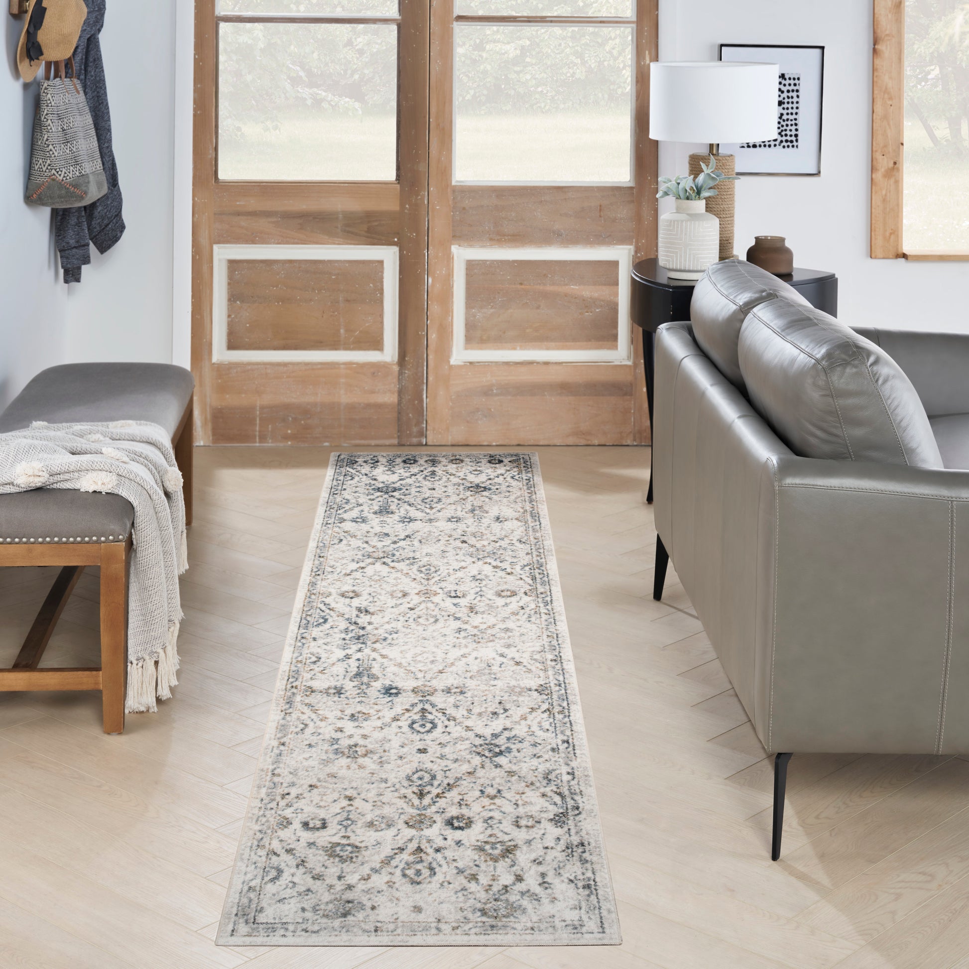 Entryway with a long gray-toned patterned runner rug, grey leather sofa, wooden doors, bench with throw blanket, and side table lamp.