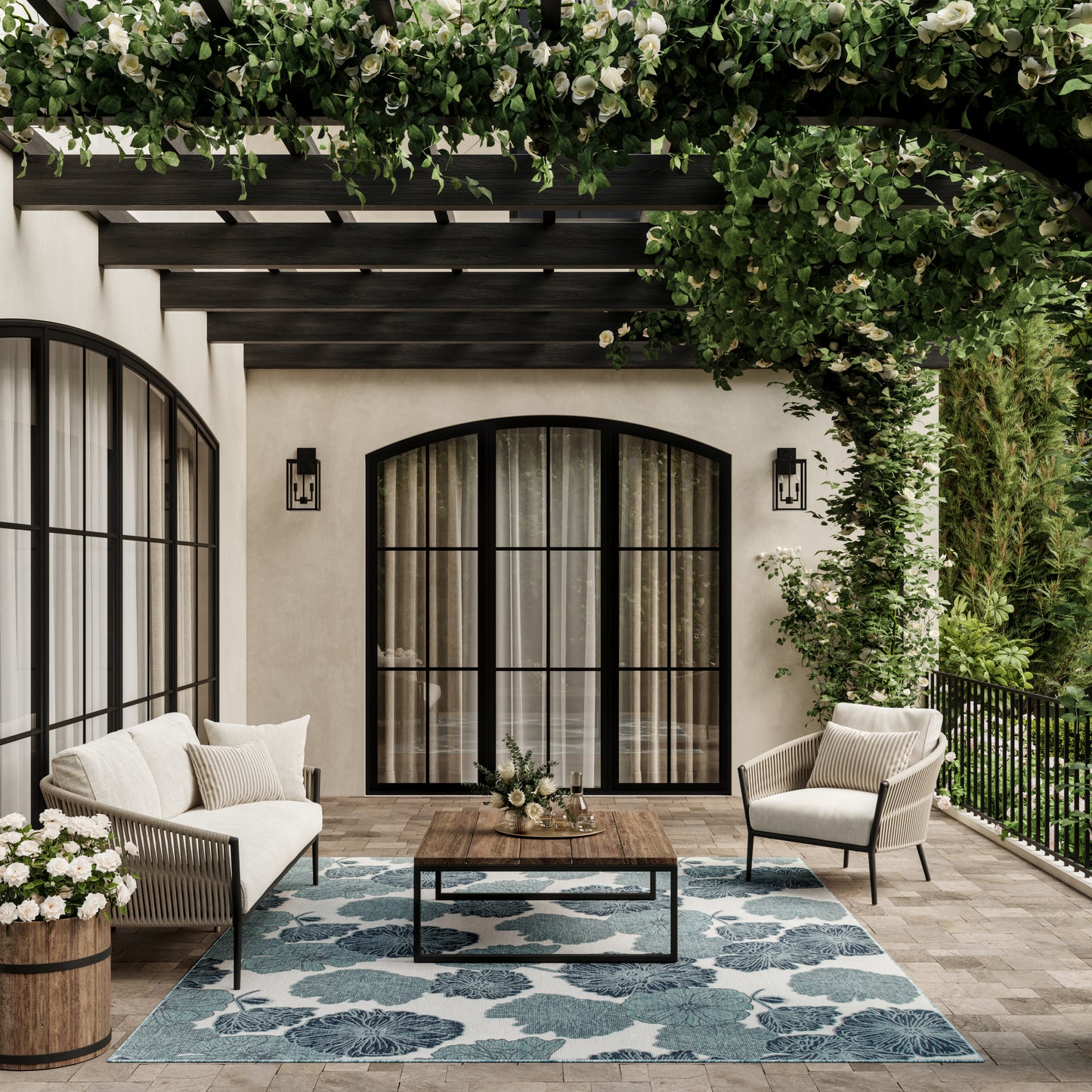 Outdoor patio with black arched glass doors, cream upholstered seating, wooden coffee table, blue floral rug, ivy pergola.
