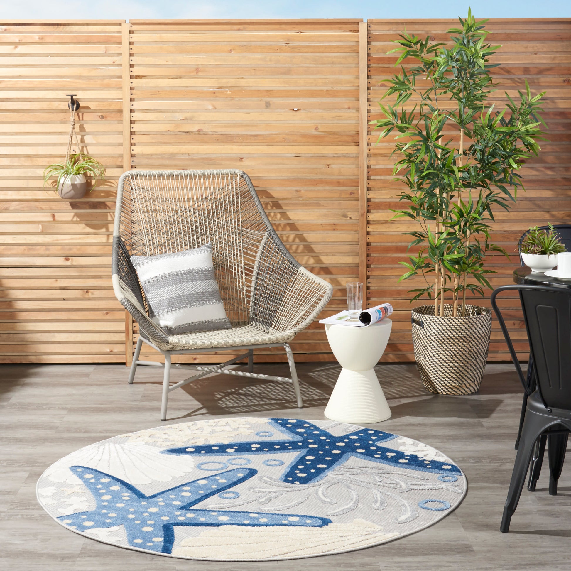 Outdoor patio scene with a woven rattan chair, gray striped cushions, round blue starfish rug, wooden privacy fence.