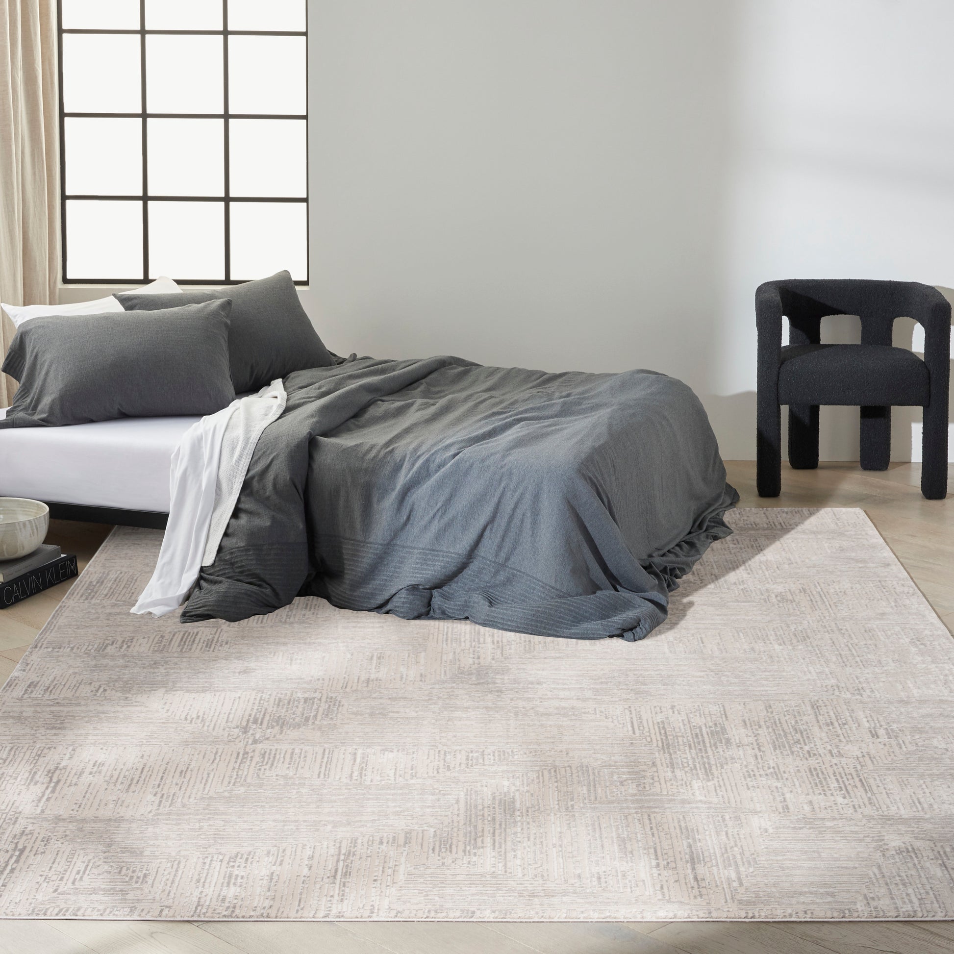 Charcoal grey bedding on a low bed with white sheets, beige rug, and a black armchair in a bright minimalist bedroom