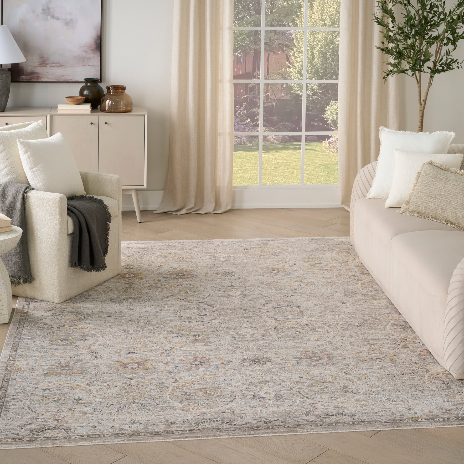 Neutral living room with beige sofa, cream armchair, patterned area rug, large window with cream curtains, and potted tree.