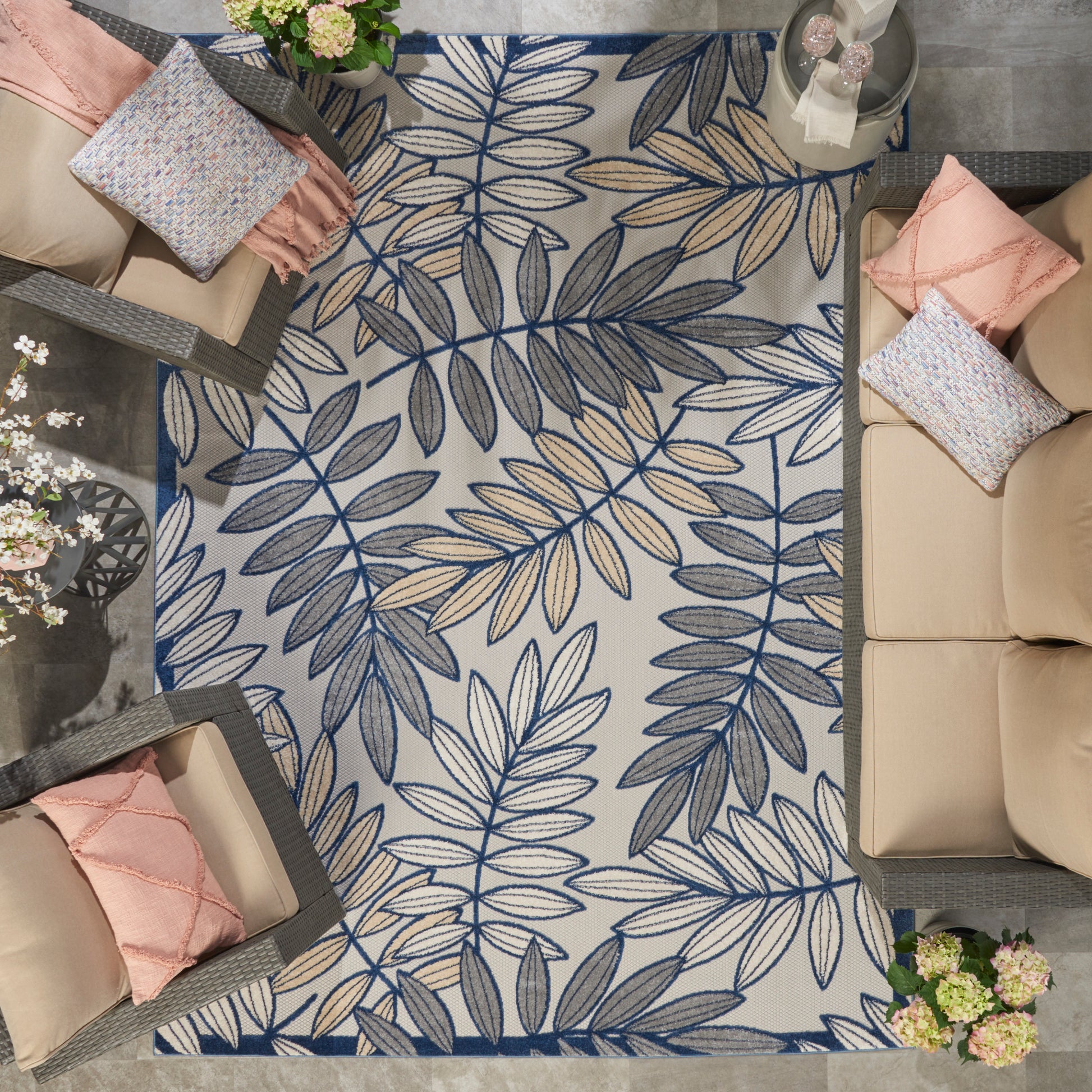 Top-view of a woven rug with a blue botanical leaf pattern on beige, surrounded by wicker sofas with pink cushions.