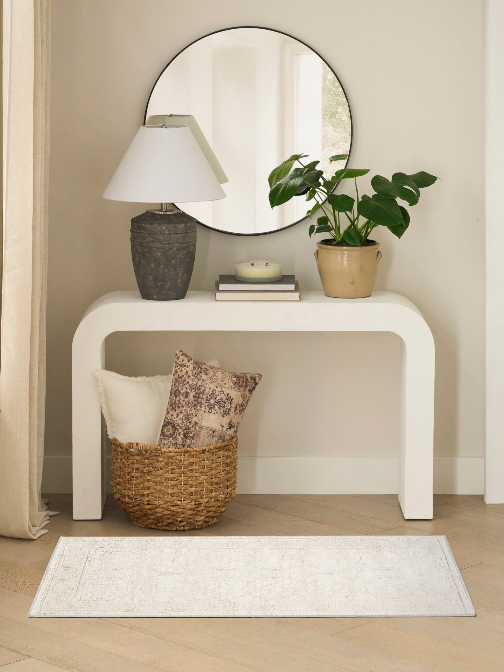 White curved console table with round mirror, stone lamp, potted plant, books and candle; wicker basket with cushions on rug.