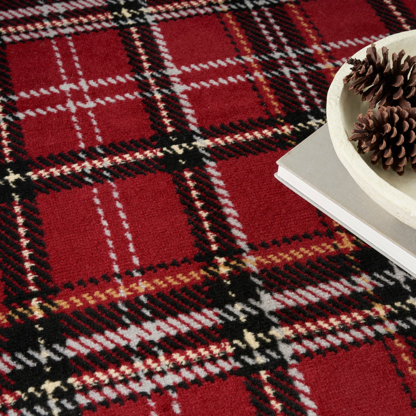 Red plaid fabric with black, white and tan lines, alongside a gray book and a white bowl holding pinecones.