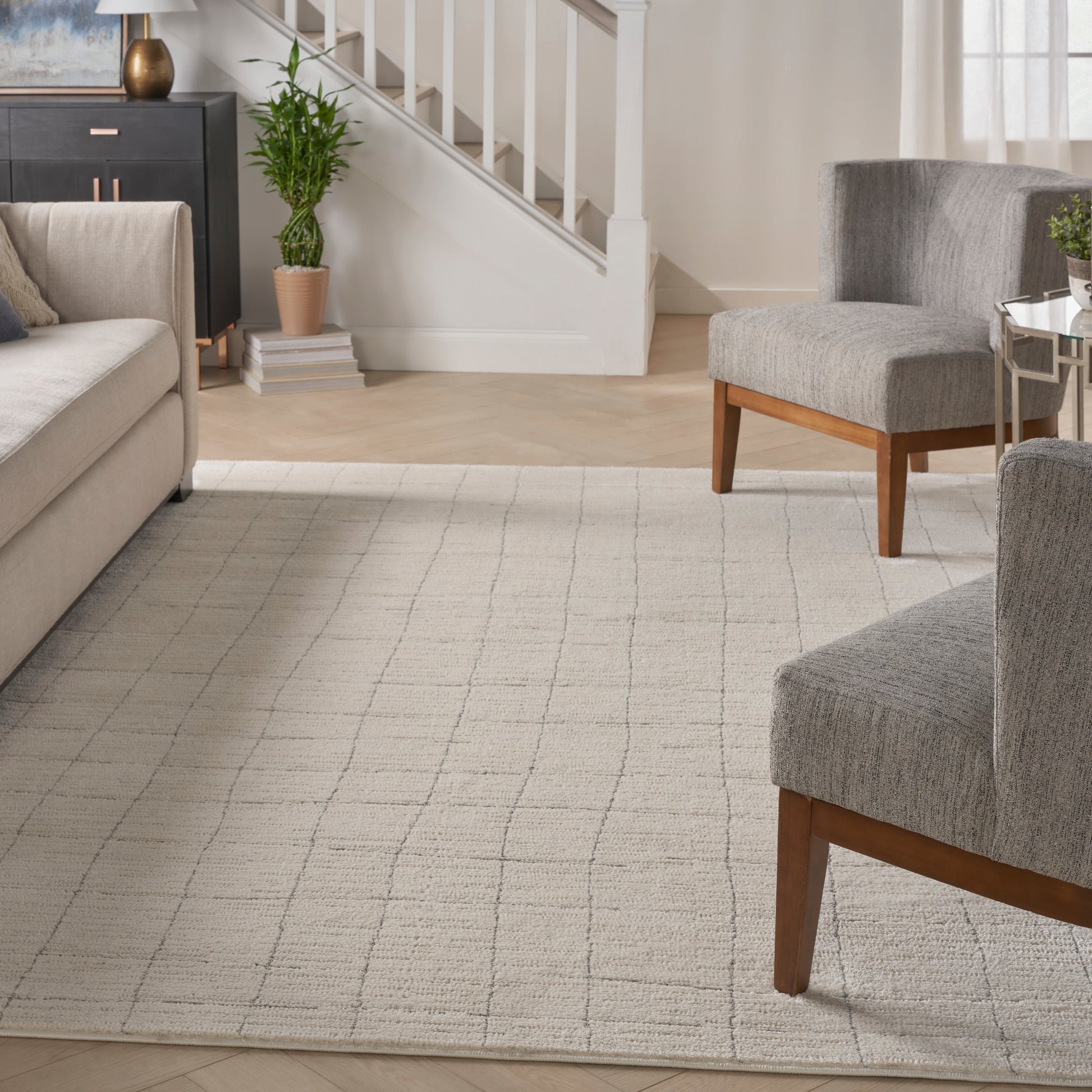Light gray grid-pattern area rug in a modern living room with gray upholstered sofa and chairs, wooden legs, staircase.