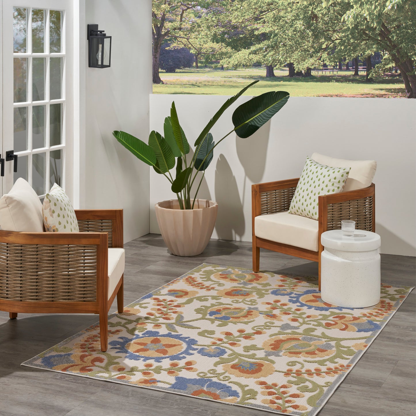 Outdoor patio with wood-framed wicker chairs, cream cushions, a large leafy plant, and a blue, orange and green floral rug.