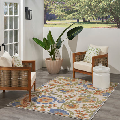 Outdoor patio with wood-framed wicker chairs, cream cushions, a large leafy plant, and a blue, orange and green floral rug.