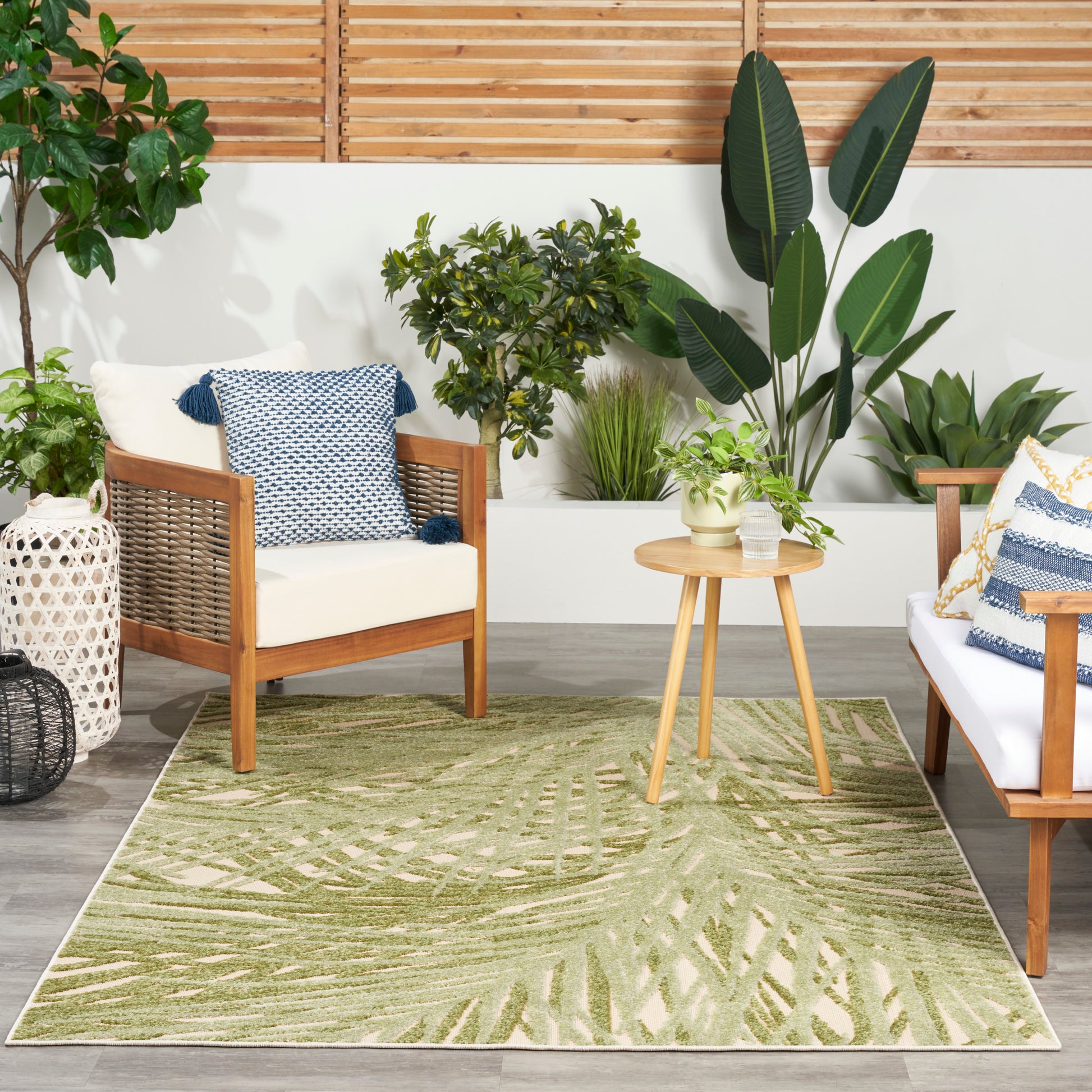 Wood-framed outdoor seating with cream cushions, blue throw pillows, round side table, and green palm-leaf rug among plants.