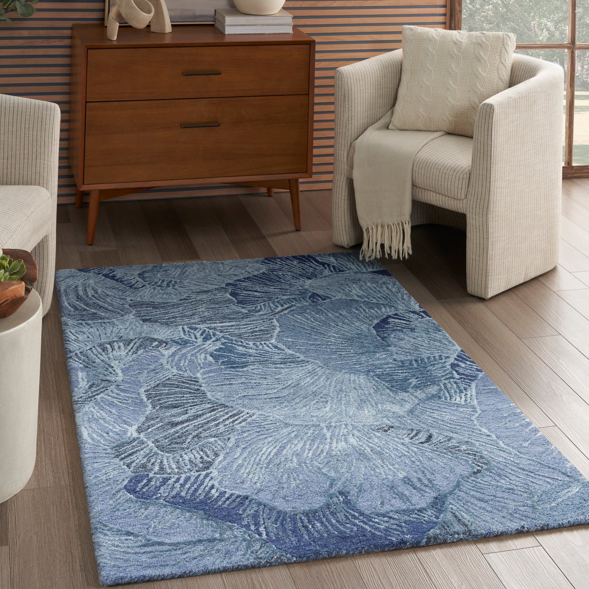 Blue rectangular area rug with pale leaf pattern in a modern living room, flanked by cream-armchairs and a wooden dresser.