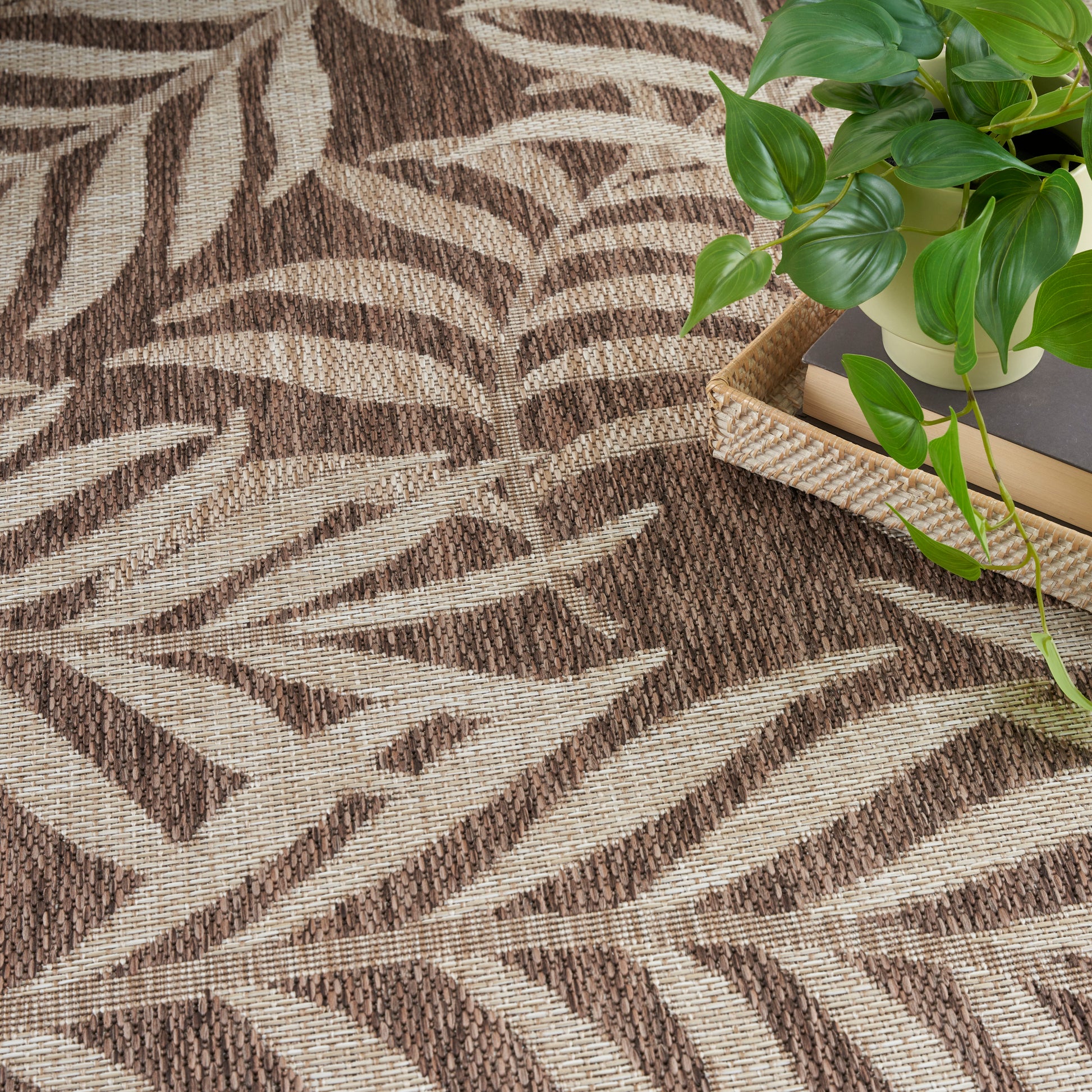 Brown and beige woven rug with leaf-like pattern, with a green pothos plant in a pot on a wicker tray with books.