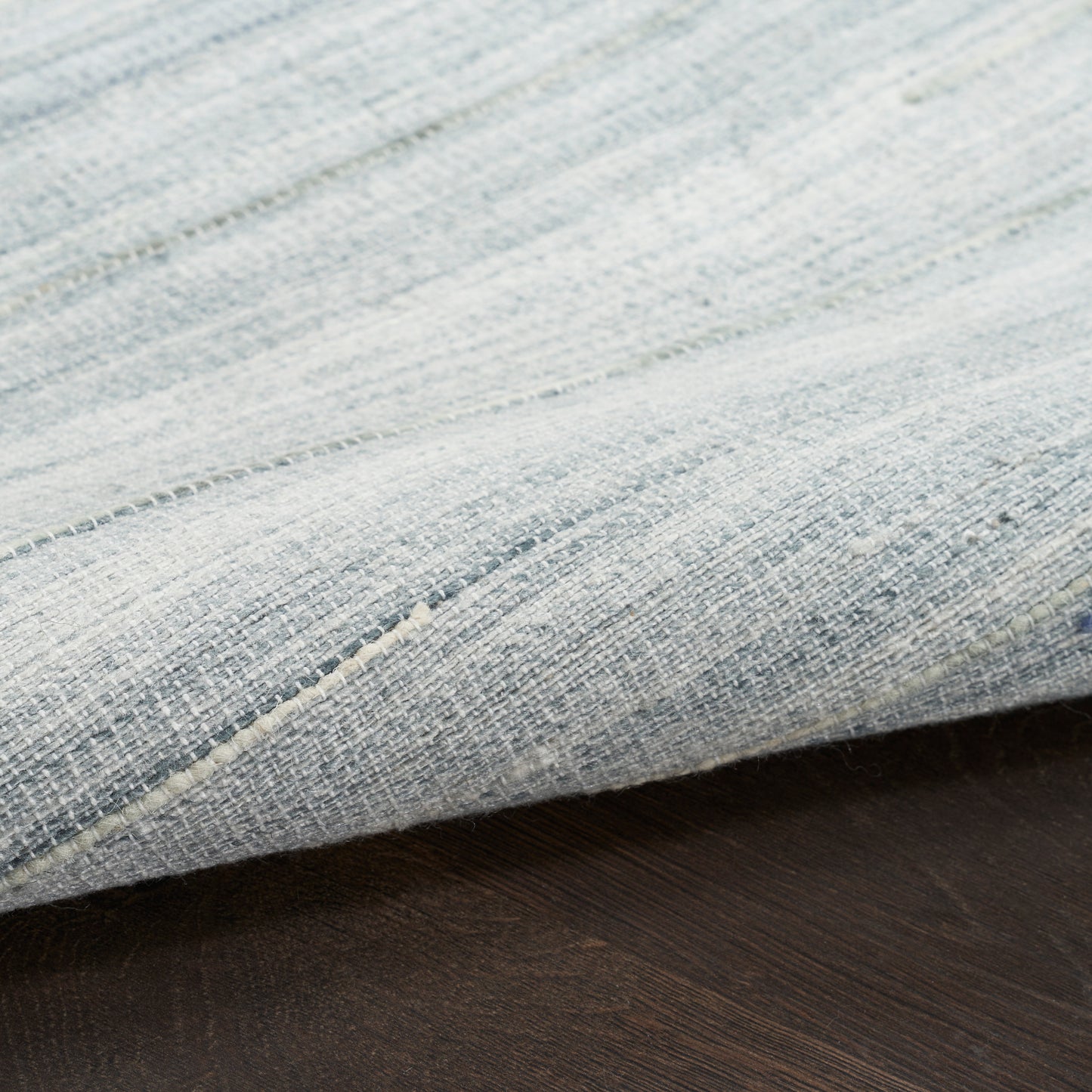 Close-up of a light blue woven rug with visible threads and a folded edge on a dark wooden floor.