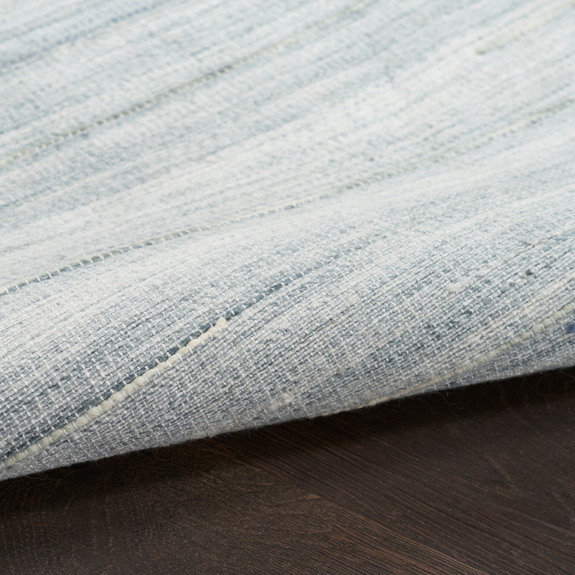 Close-up of a light blue woven rug with visible threads and a folded edge on a dark wooden floor.