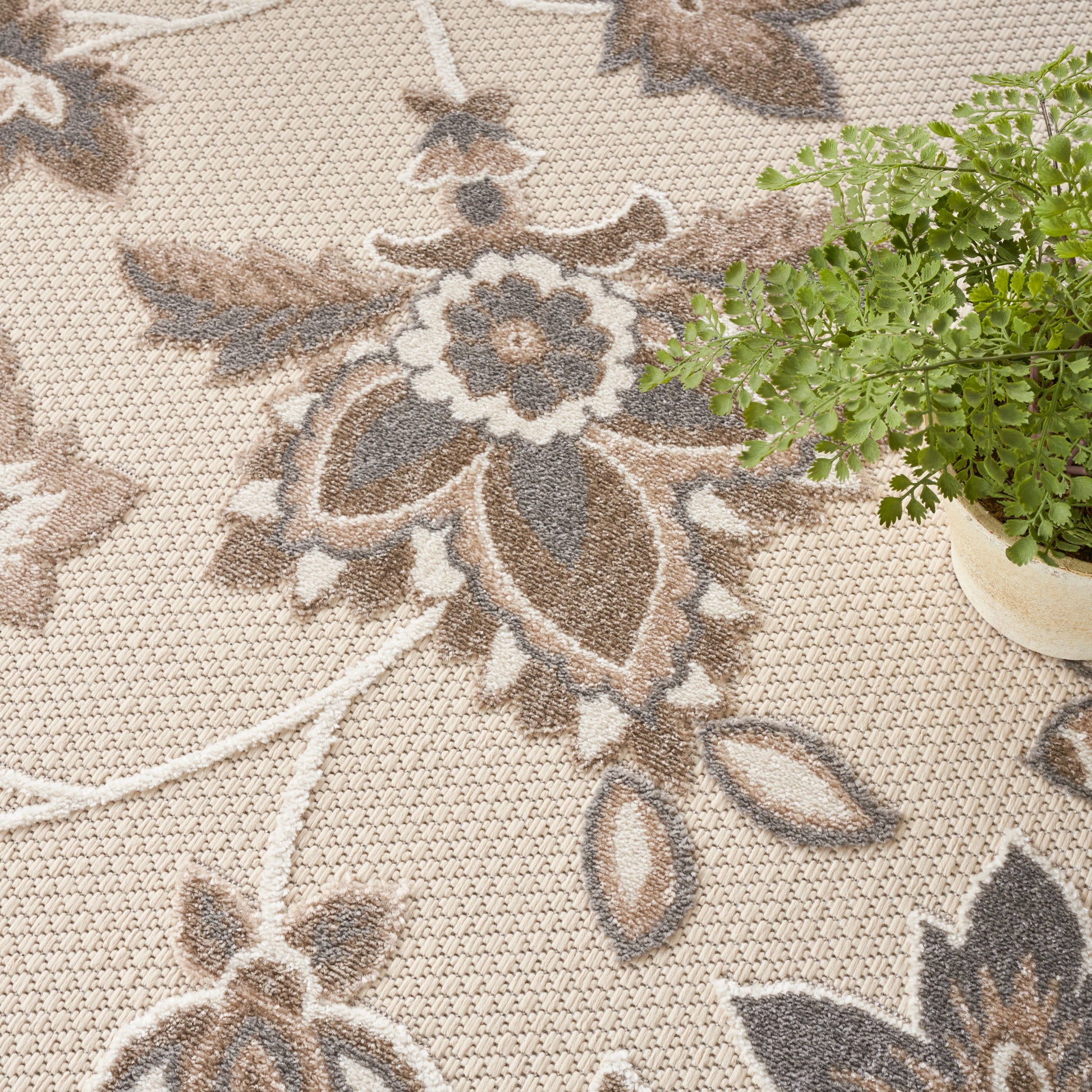 Beige floral patterned rug with brown and gray motifs; textured woven surface, paired with a small potted fern
