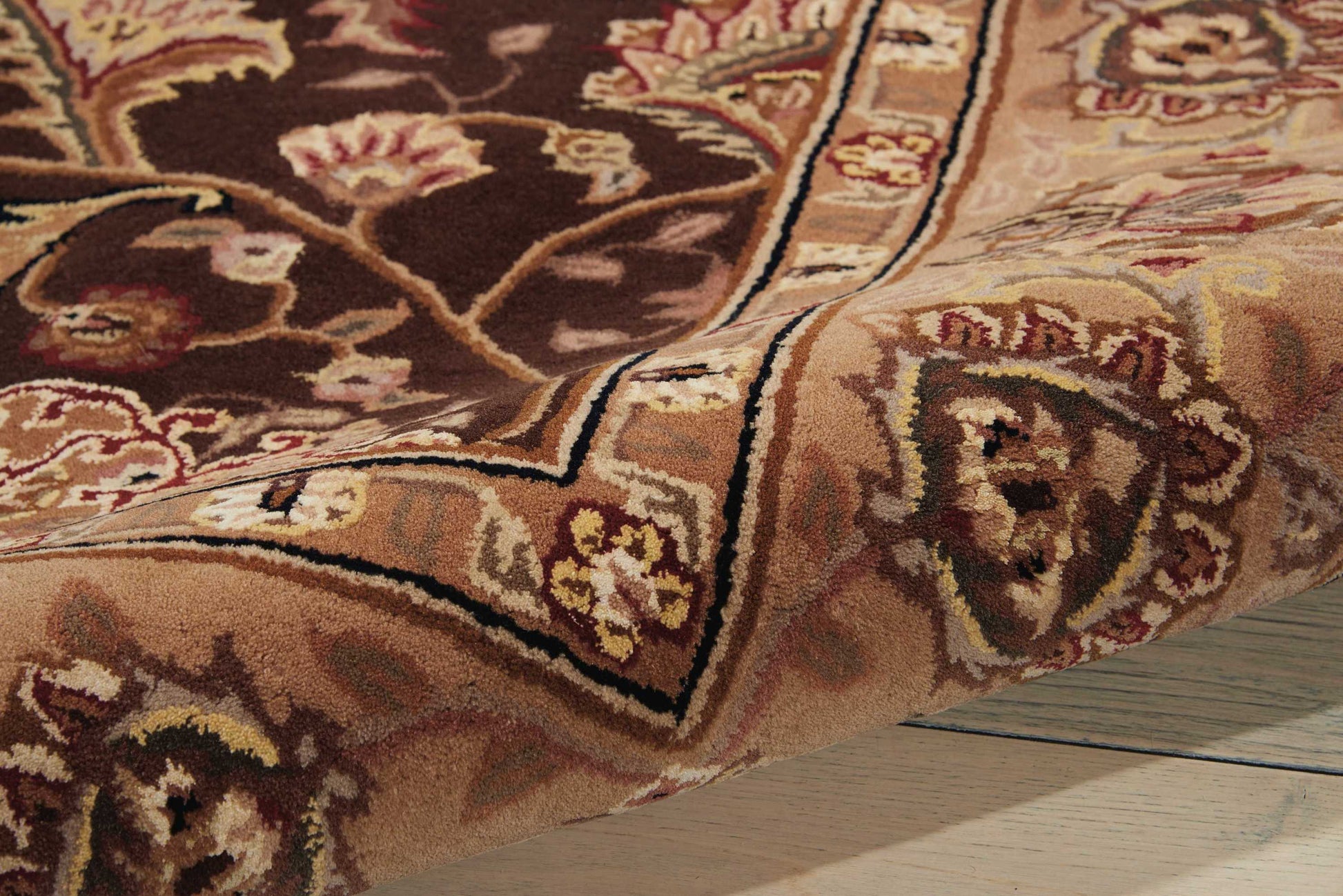 Close-up of an ornate oriental rug with brown, tan, cream and burgundy floral pattern, folded on a wooden floor.