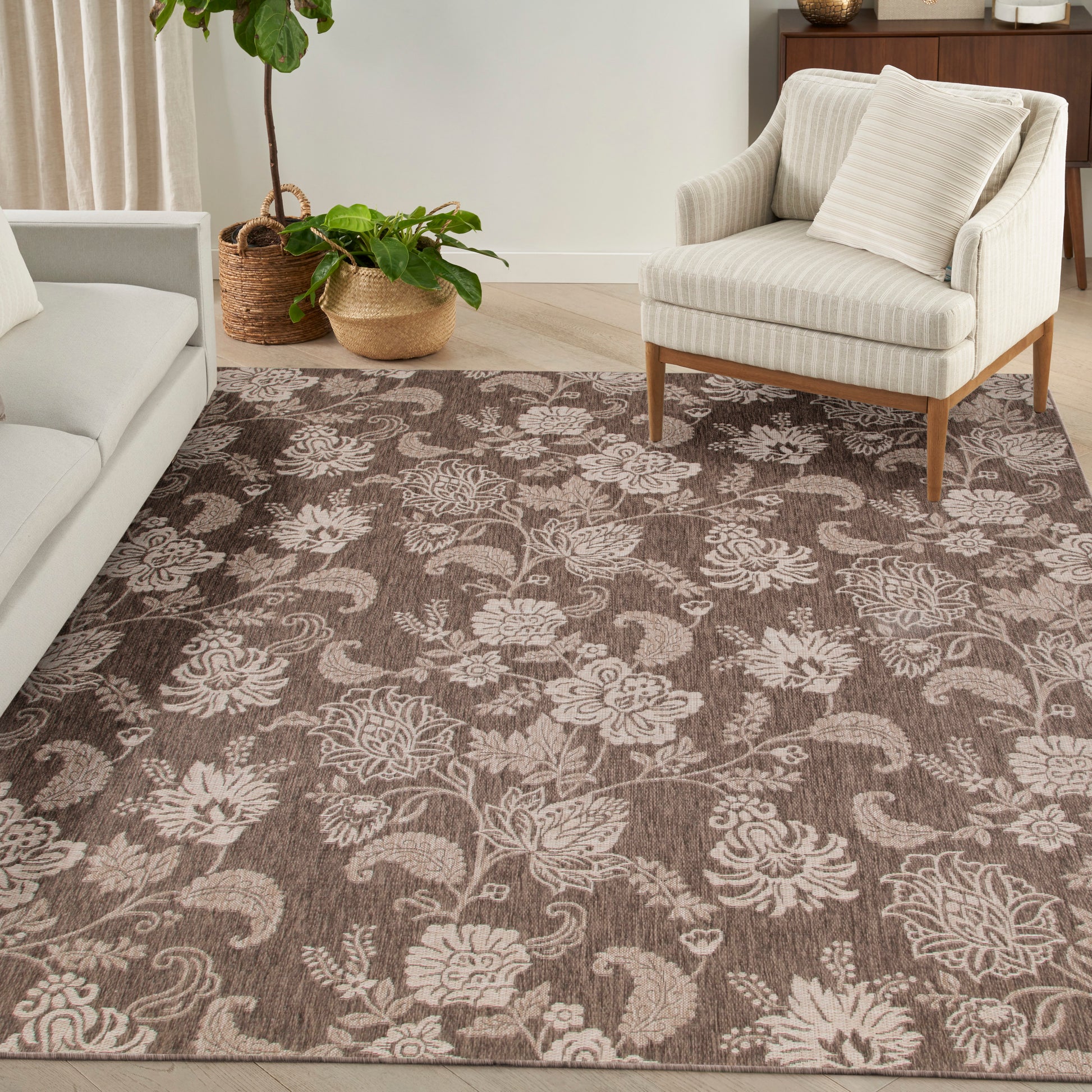 Brown floral area rug with cream botanical pattern in a neutral living room with a beige upholstered sofa and wooden-legged armchair.
