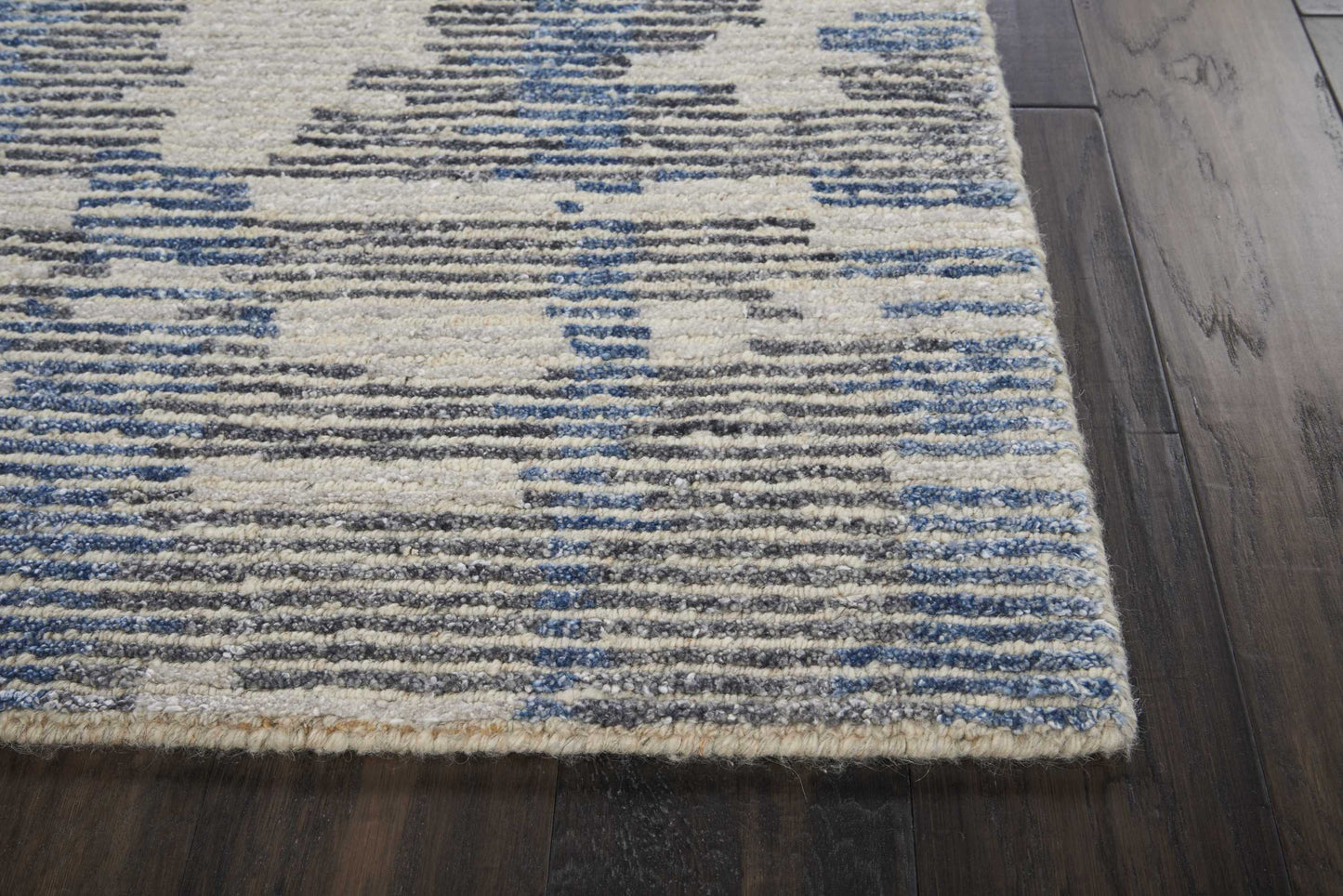 Beige area rug with blue and gray geometric pattern on dark wood floor, woven texture, short fringe along edge.