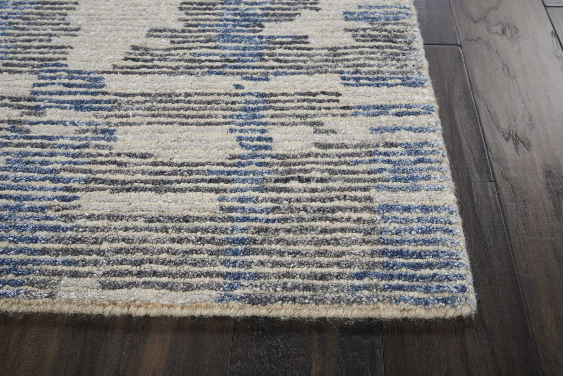 Beige area rug with blue and gray geometric pattern on dark wood floor, woven texture, short fringe along edge.