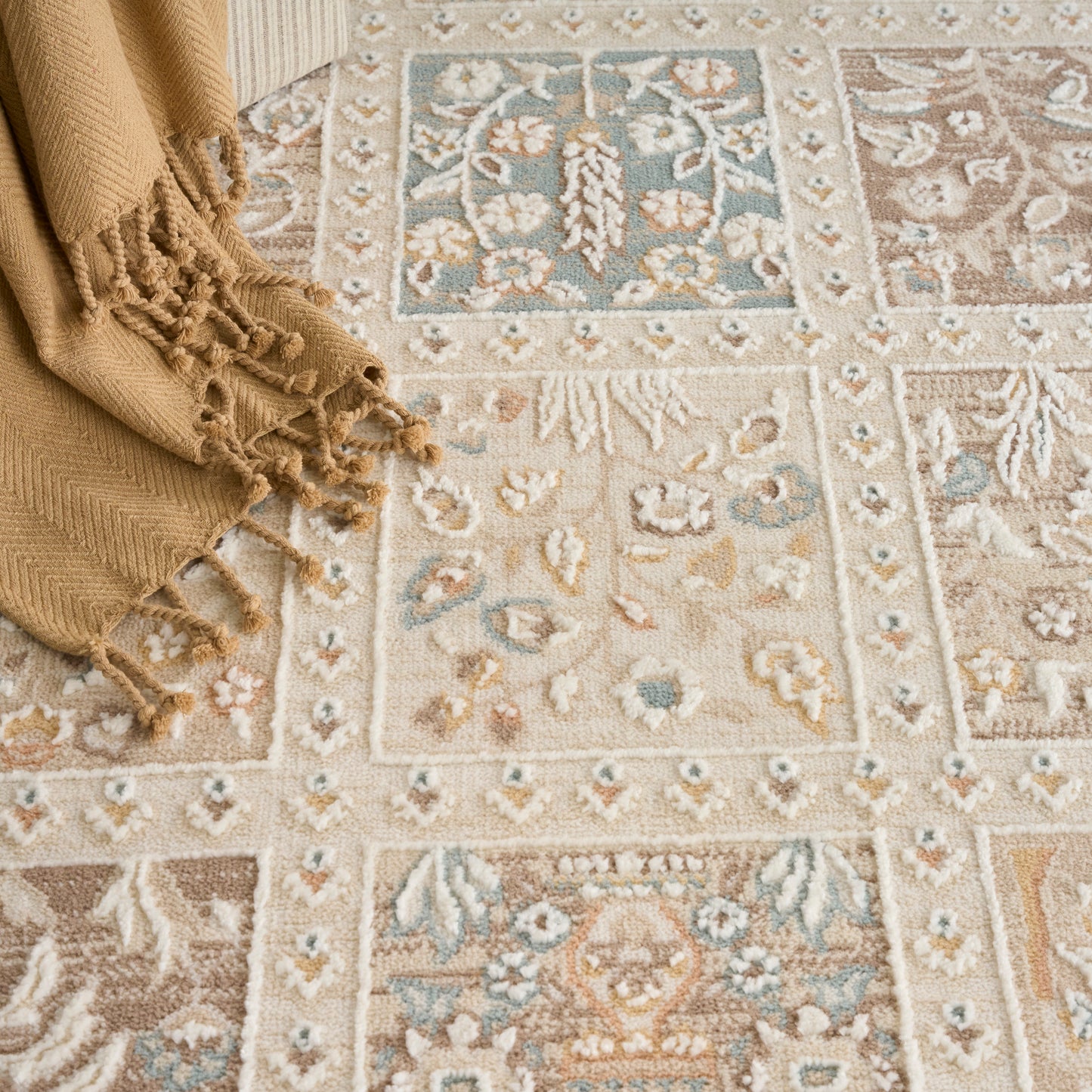 Beige oriental rug with blue, brown and cream floral medallions and borders; tan fringed throw draped at corner.