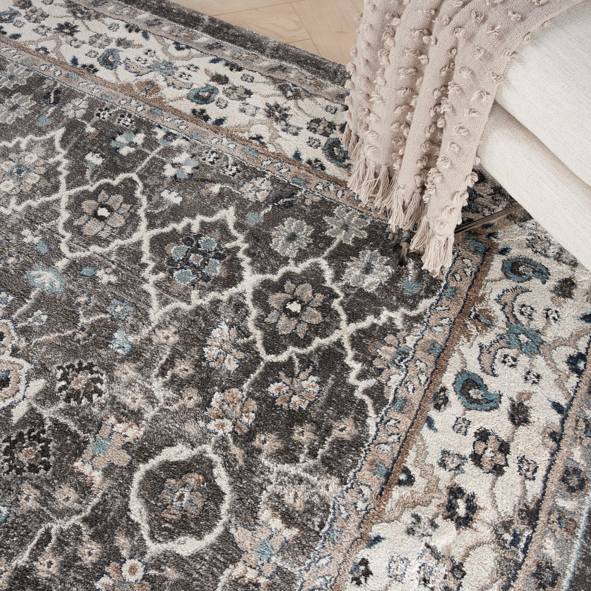 Gray ornate area rug with blue floral motifs and beige border, draped pale pink blanket with fringe.