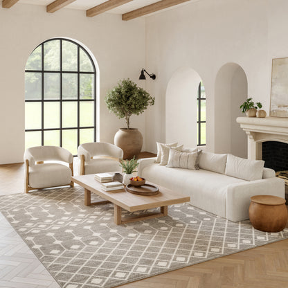 Neutral living room with arched window, beige sofa, cream armchairs, wooden coffee table, patterned rug, tree, fireplace.