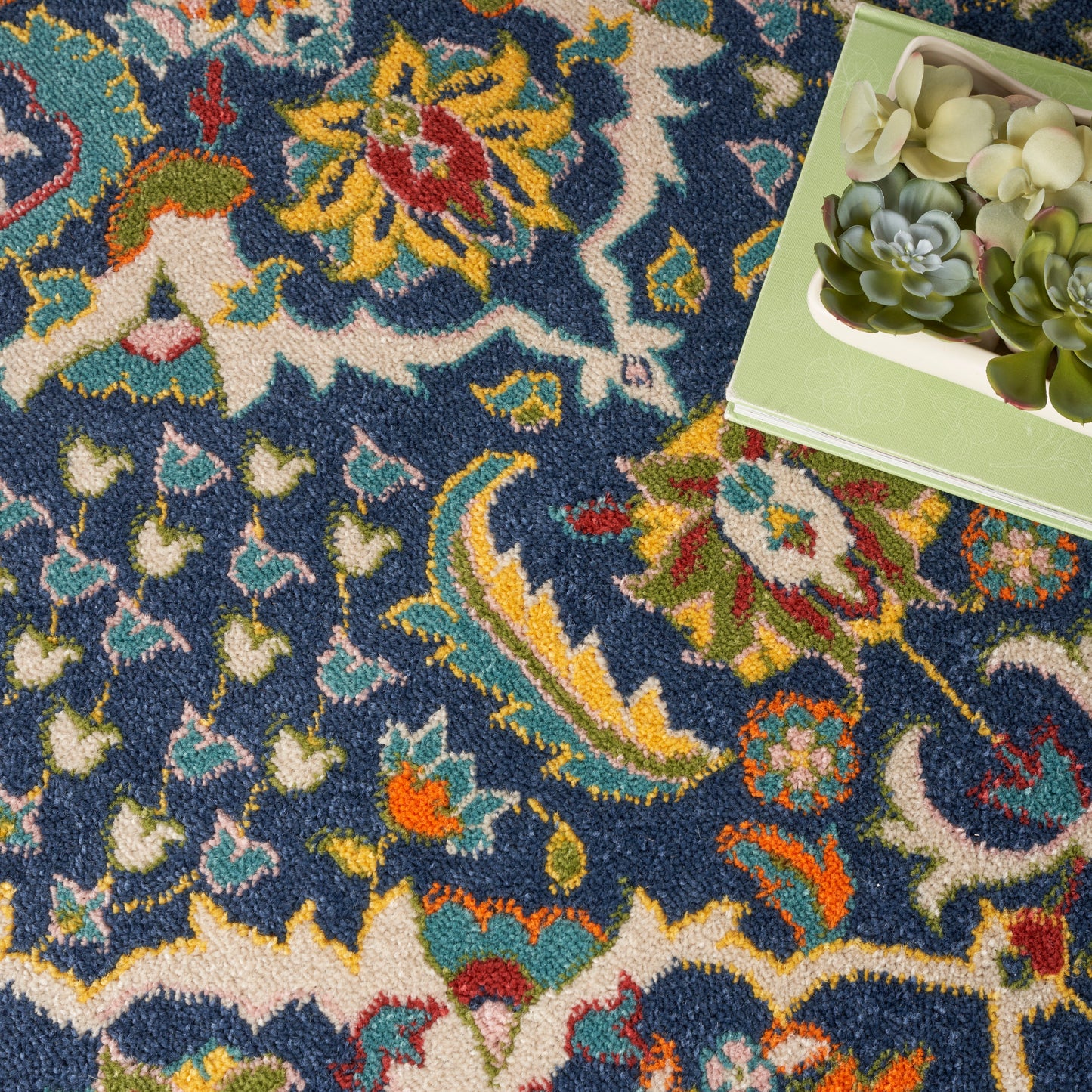 Colorful Persian-style rug with blue field & yellow, red, teal florals; green succulent box on top-right