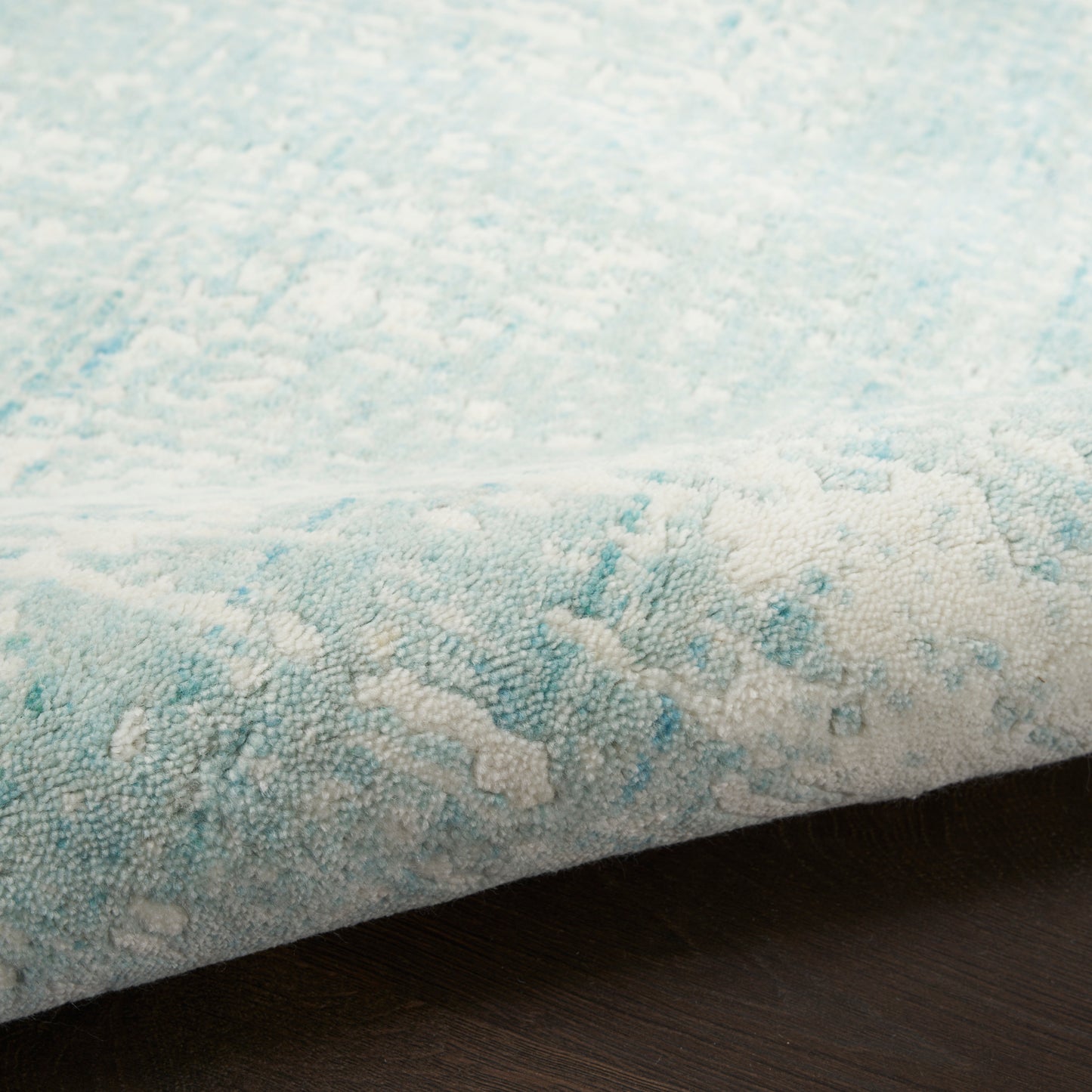 Light blue and white textured rug with subtle abstract pattern, edge slightly curled on dark wood floor.
