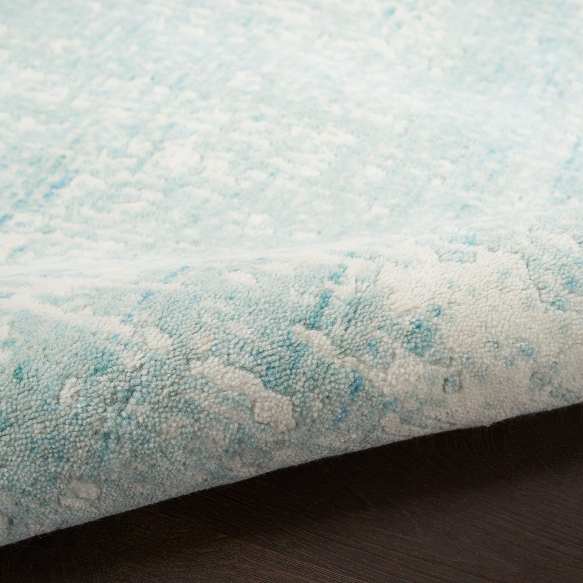 Light blue and white textured rug with subtle abstract pattern, edge slightly curled on dark wood floor.
