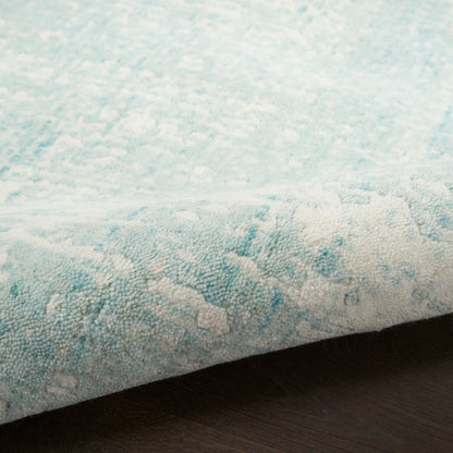 Light blue and white textured rug with subtle abstract pattern, edge slightly curled on dark wood floor.