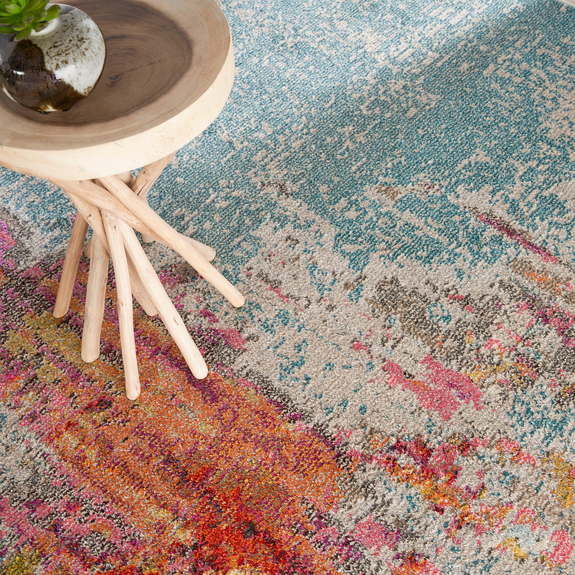 Vibrant multicolor abstract patterned rug with a light wood round-top stool with spindle legs and a ceramic pot with a plant on top of the stool.