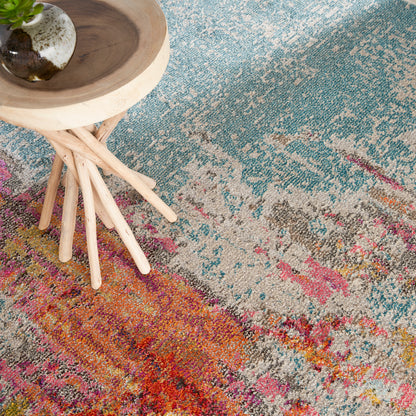 Vibrant multicolor abstract patterned rug with a light wood round-top stool with spindle legs and a ceramic pot with a plant on top of the stool.
