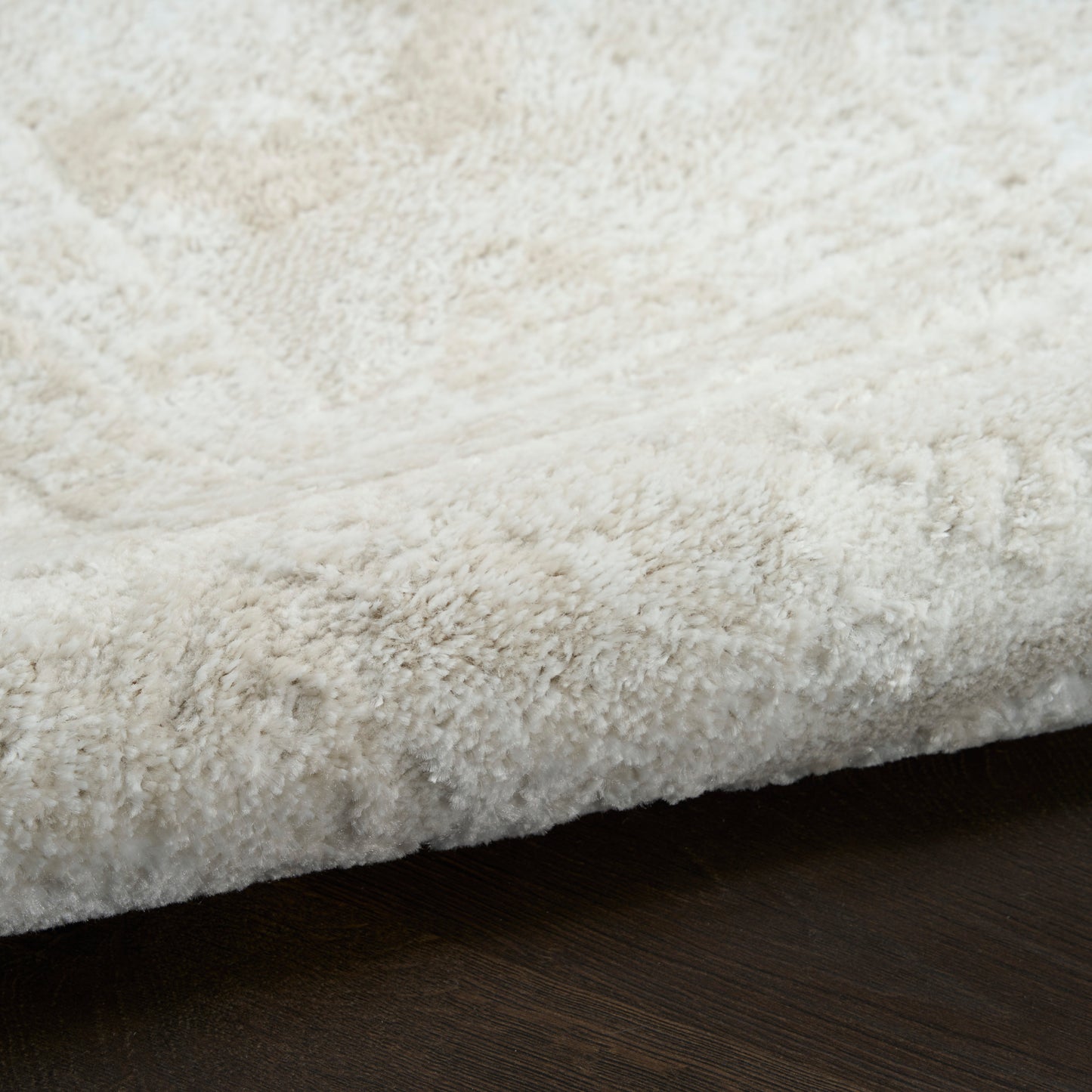 Cream shag rug with dense fluffy pile resting on dark wood floor, close-up edge view.