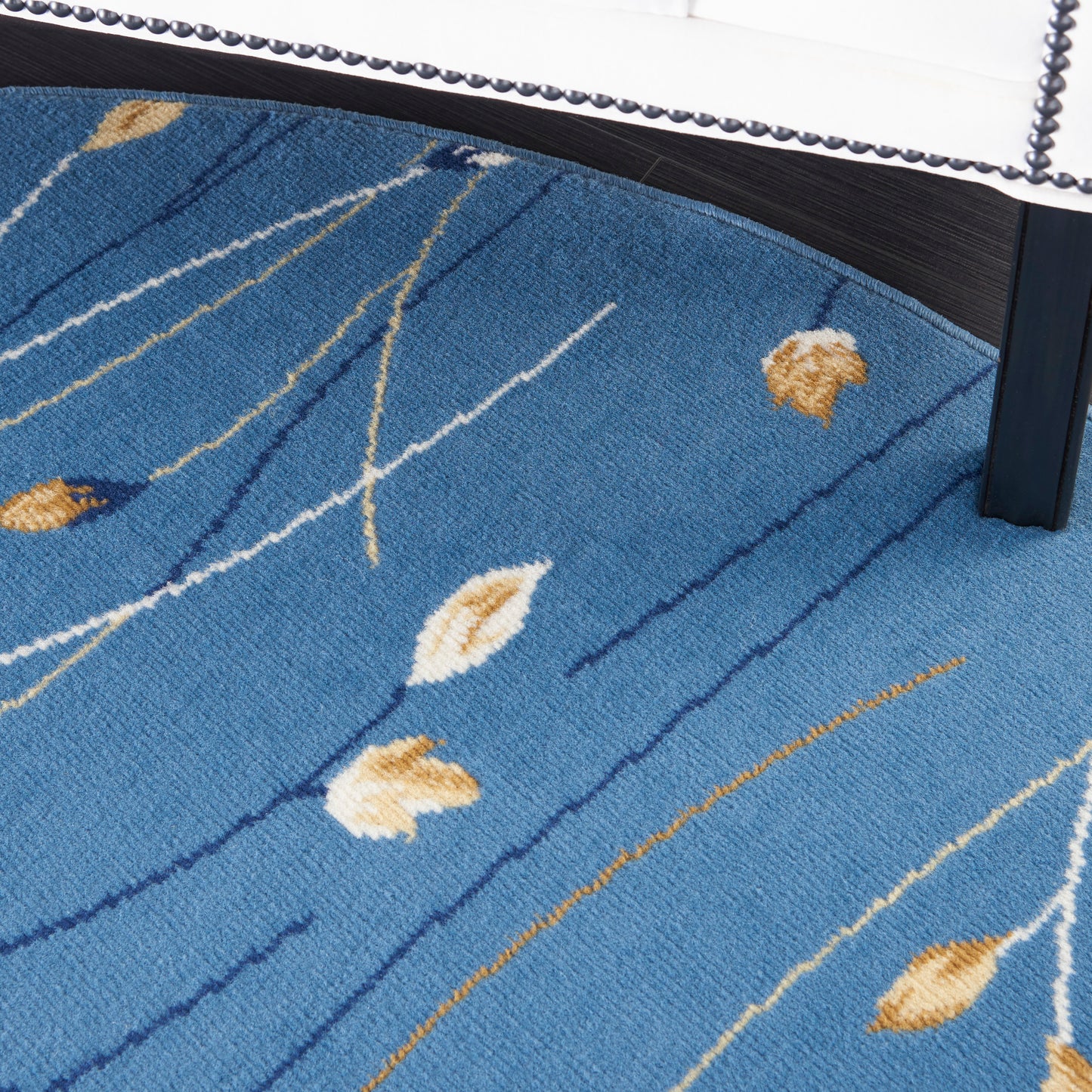 Blue textured rug with white and gold leaf shapes and curved lines under a white upholstered bed with nailhead trim