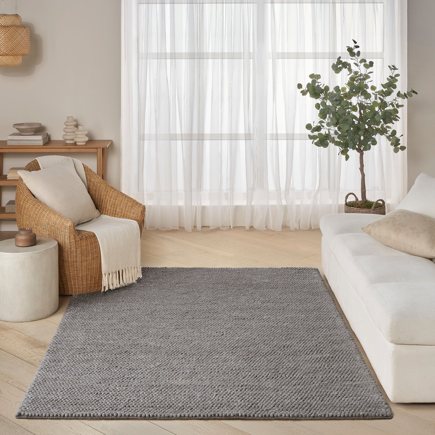Cozy living room with light wood floor, grey woven rug, sofa, beige wicker chair with throw, potted tree, sheer curtains.