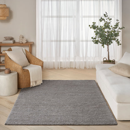 Cozy living room with light wood floor, grey woven rug, sofa, beige wicker chair with throw, potted tree, sheer curtains.