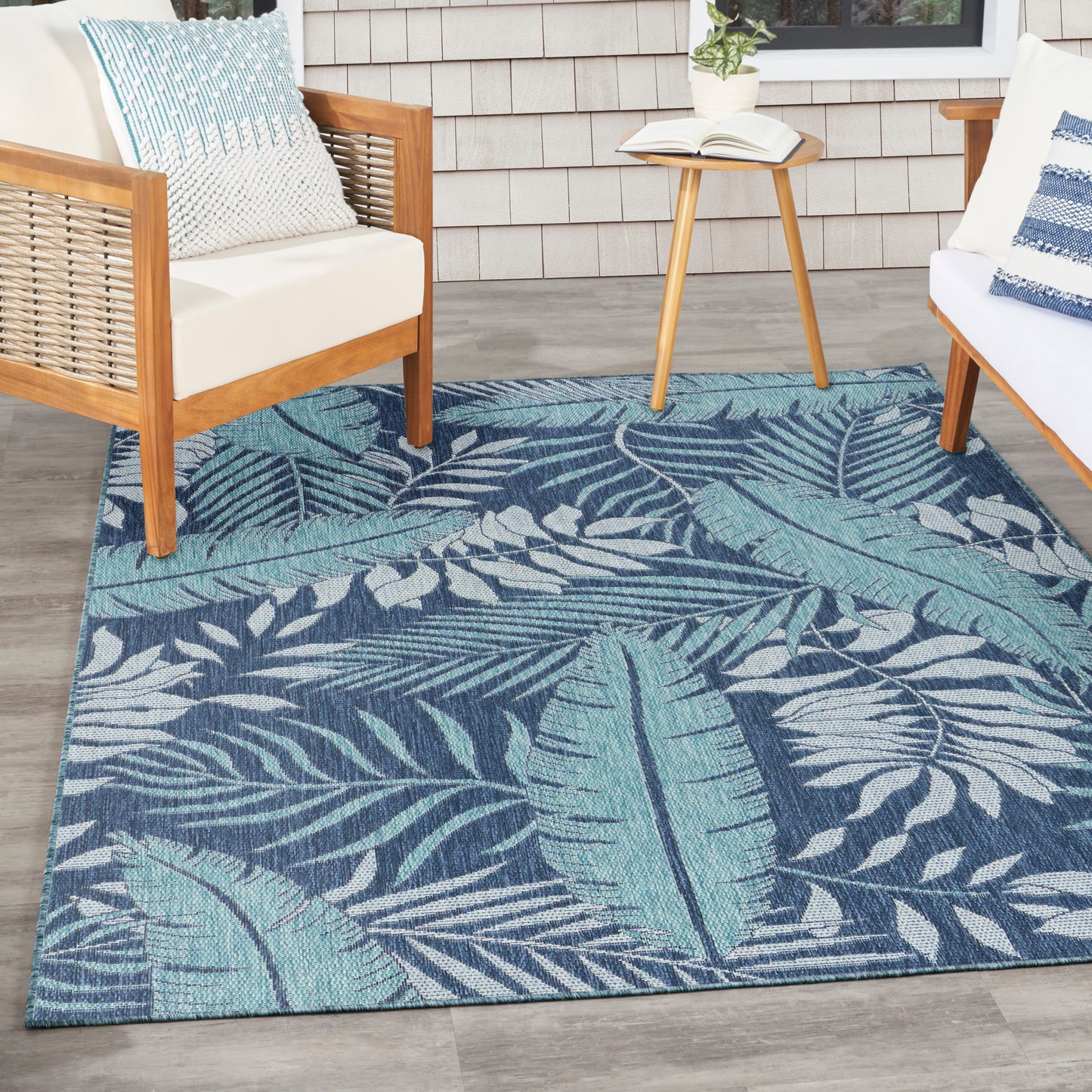 Rectangular teal-blue tropical palm leaf outdoor rug on patio with wooden-framed seating and a round side table.