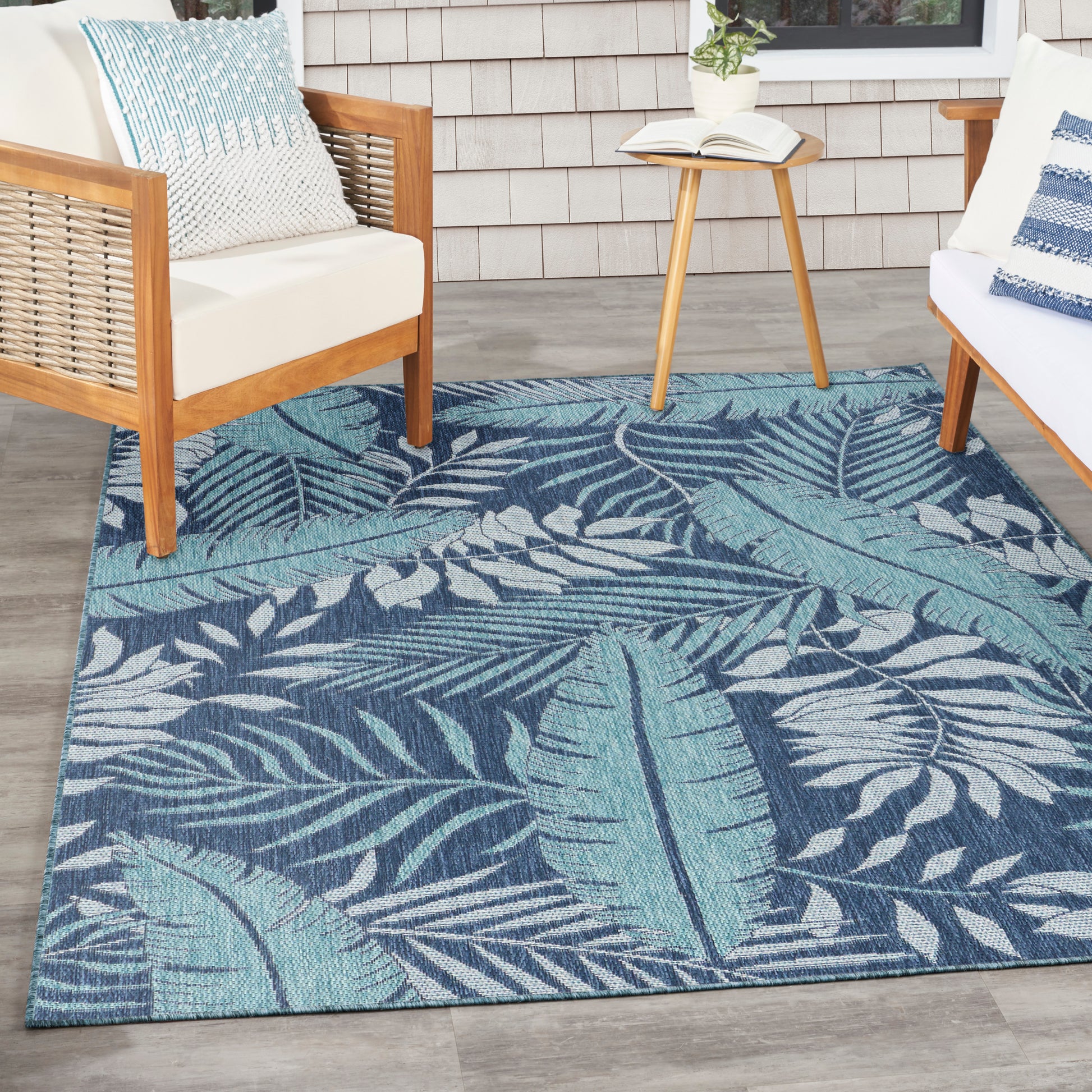 Rectangular teal-blue tropical palm leaf outdoor rug on patio with wooden-framed seating and a round side table.