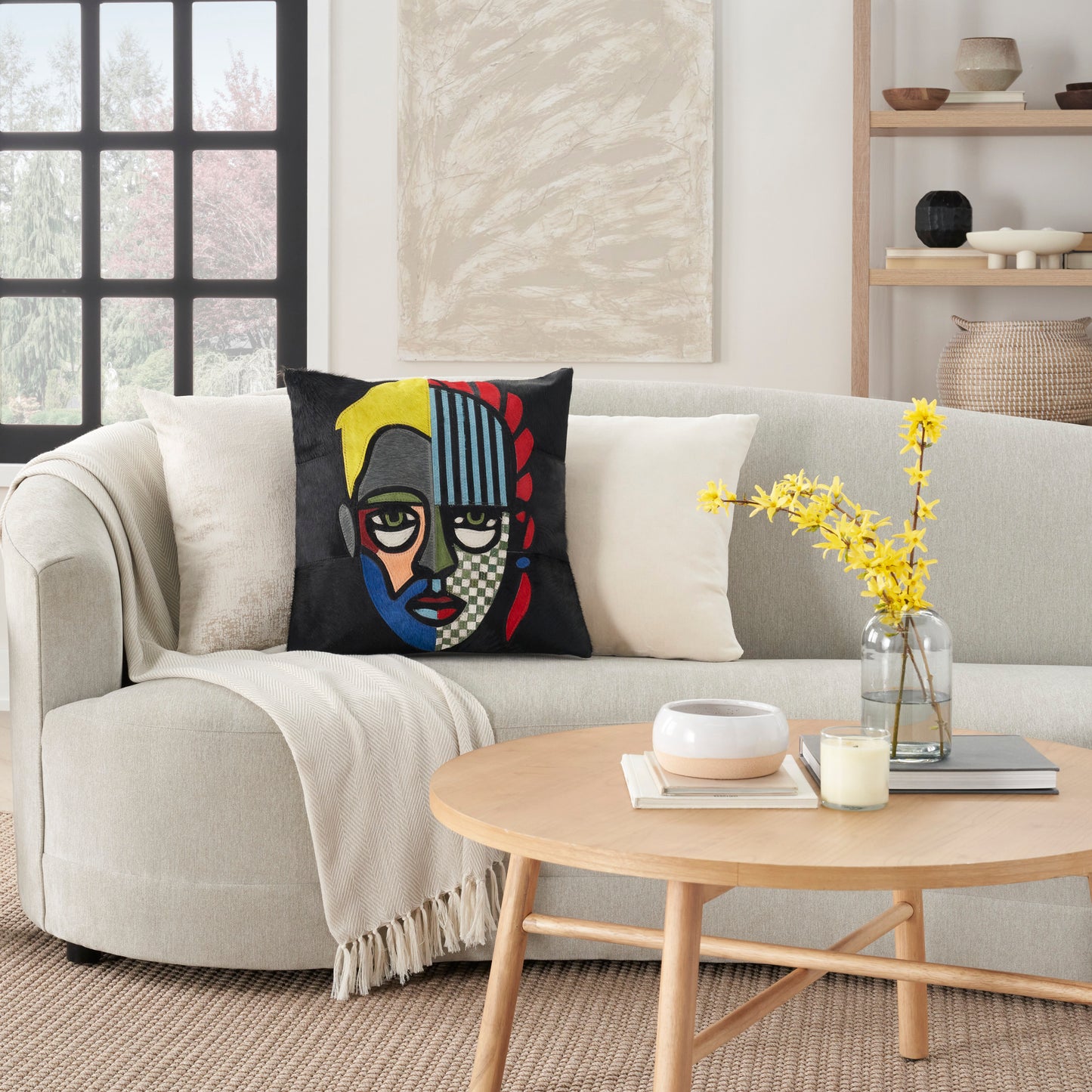 Beige sofa with cream throw & bold abstract pillow; round wood coffee table, yellow flowers, candles, & books.