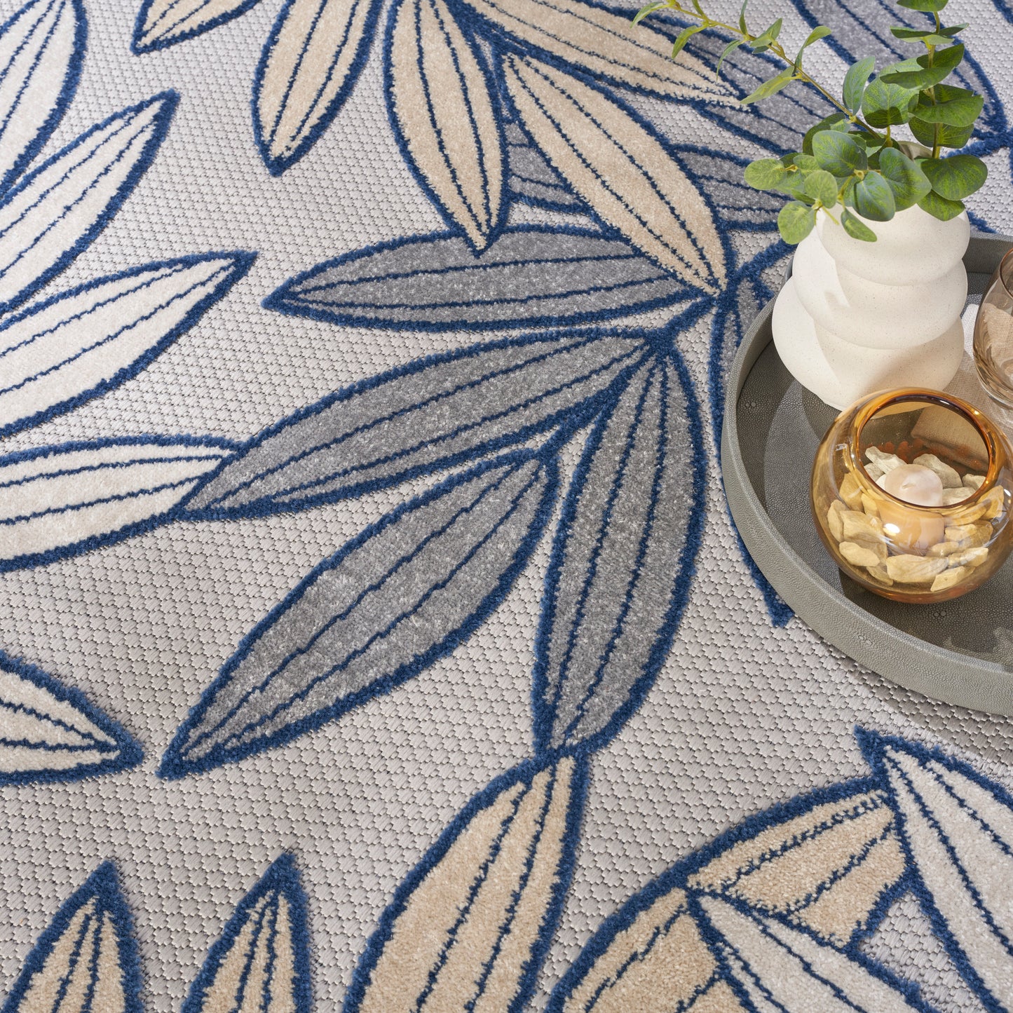 Beige rug with dark blue leaf pattern and textured weave; plant and candle on a gray round tray beside.